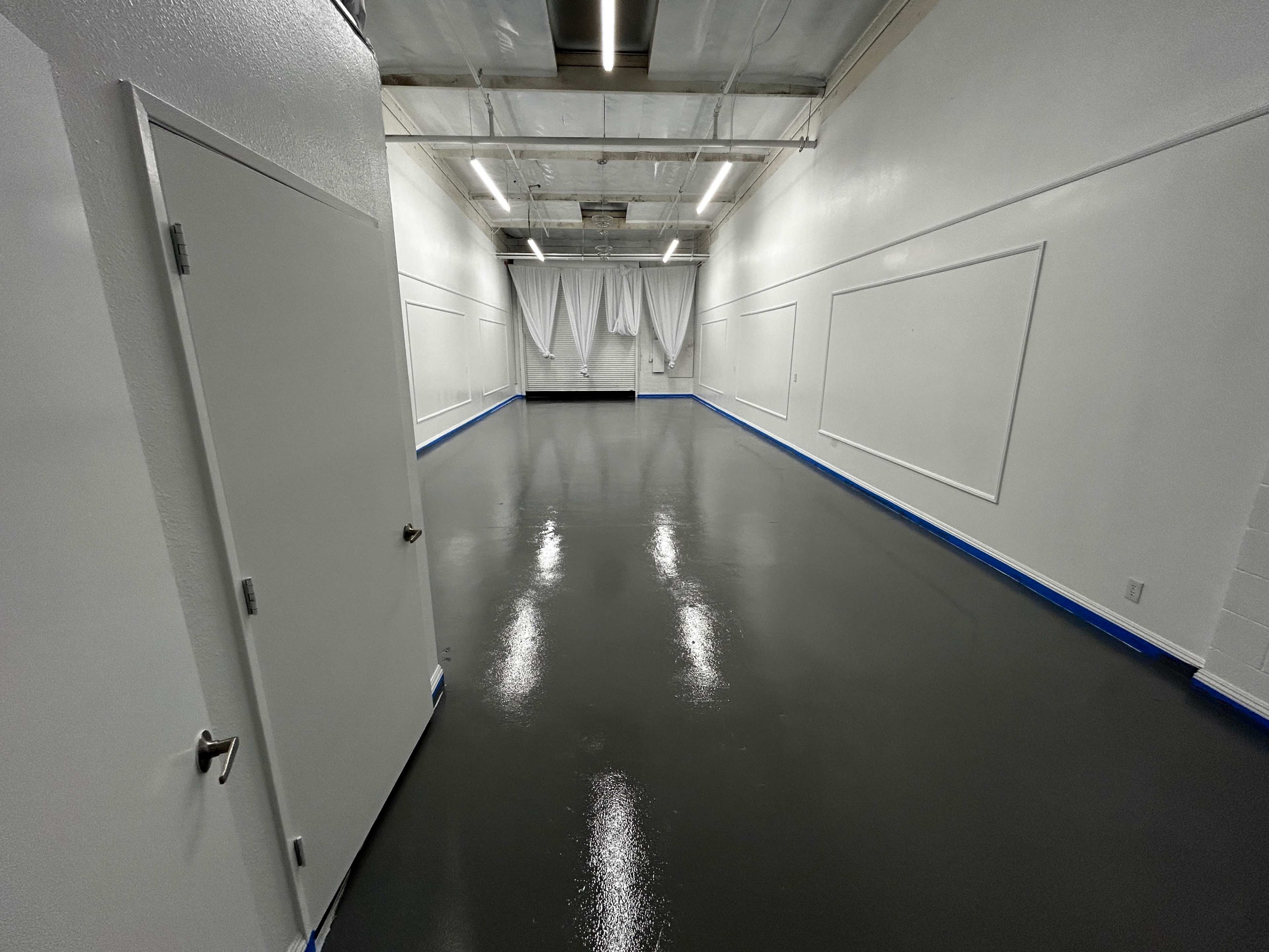 A spacious, empty room with a reflective gray floor and white walls, featuring a closed door on one side and a curtain at the far end.