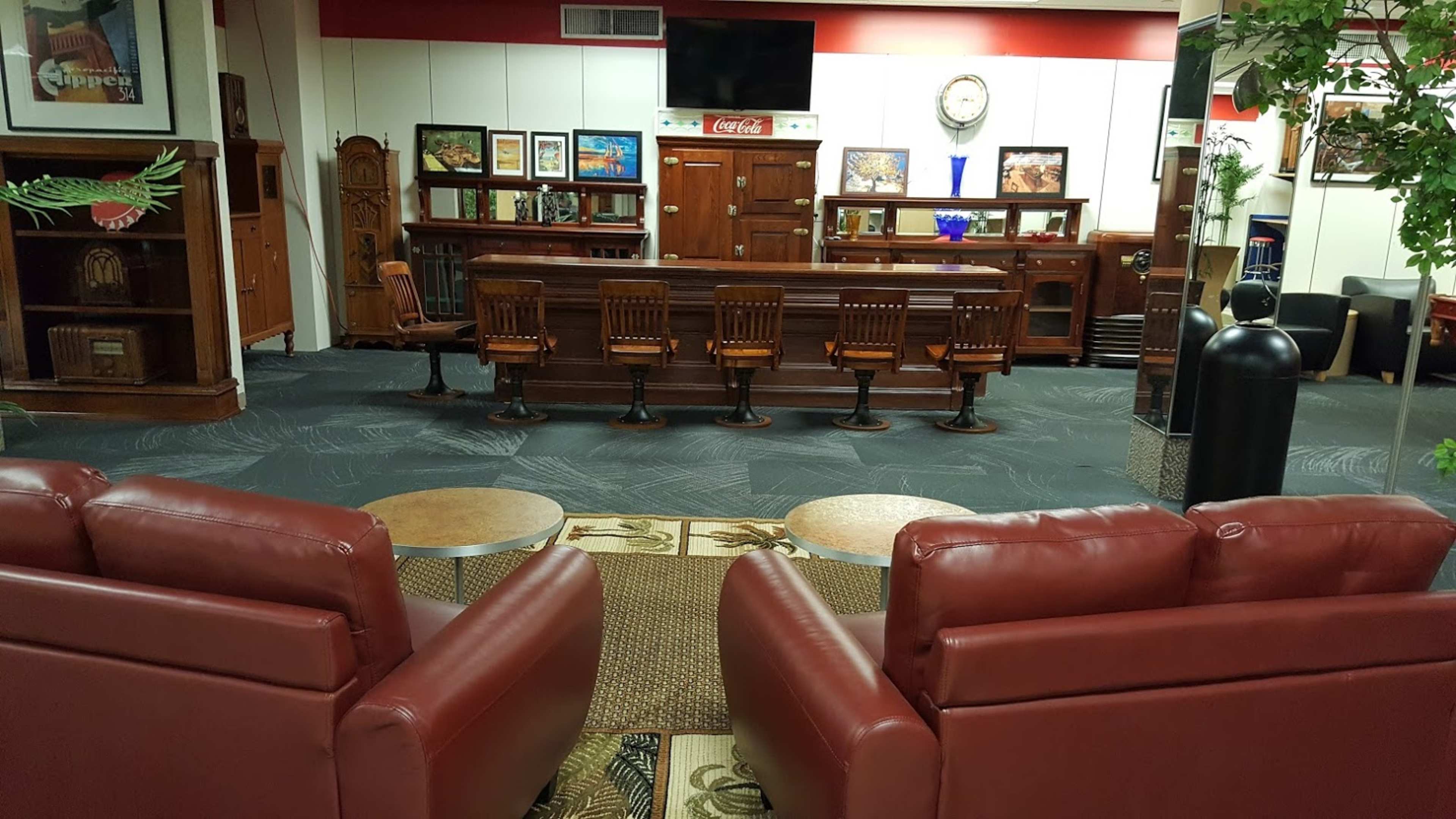 A lounge area with two red chairs facing a wooden bar and a row of high chairs, surrounded by decorative elements and framed pictures on the walls.