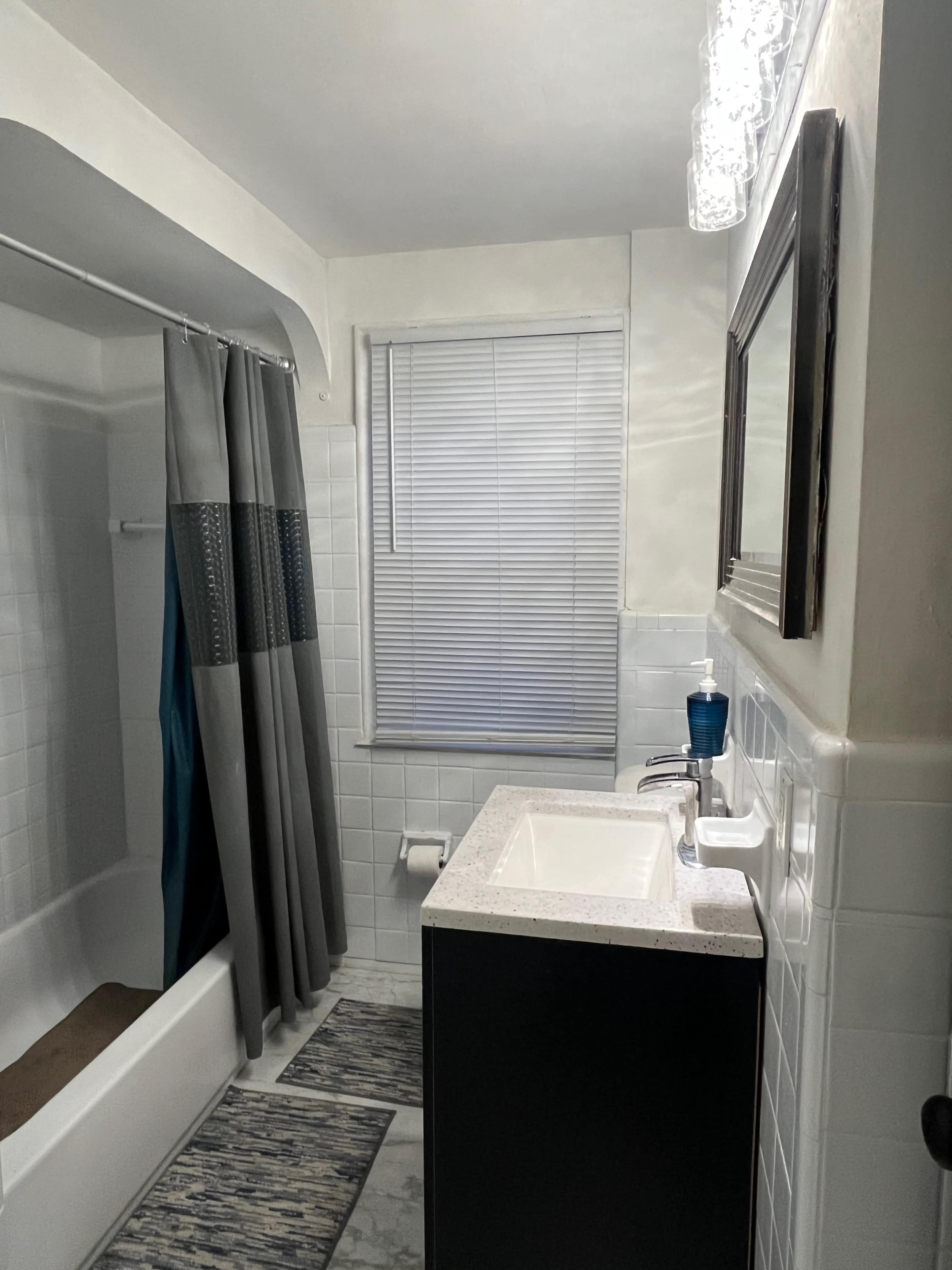 The image shows a small bathroom with a bathtub, a gray shower curtain, a white sink with a dark cabinet, and a window with blinds.