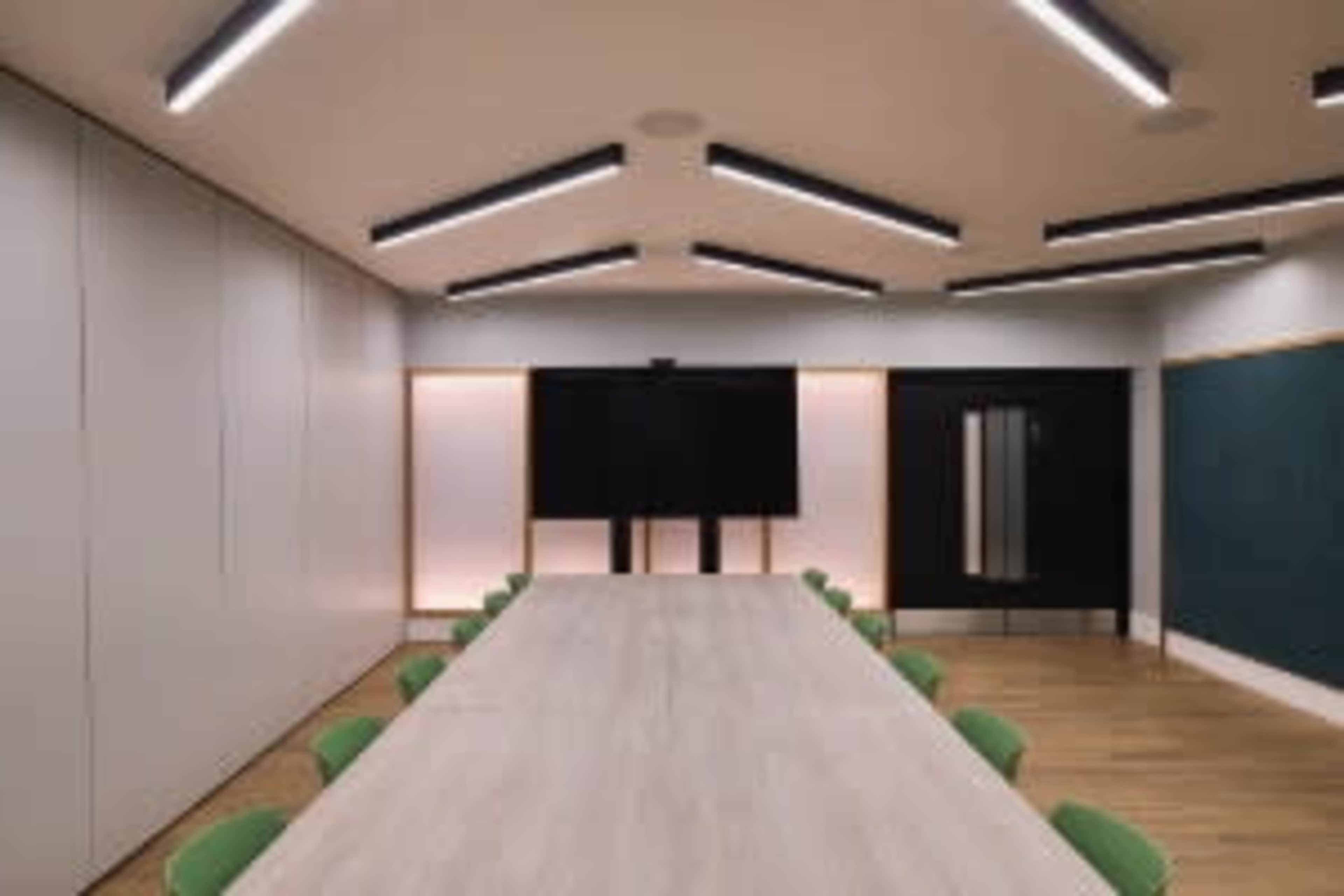 A spacious conference room features a long wooden table surrounded by green chairs, with a large screen and a black-paneled wall at one end.