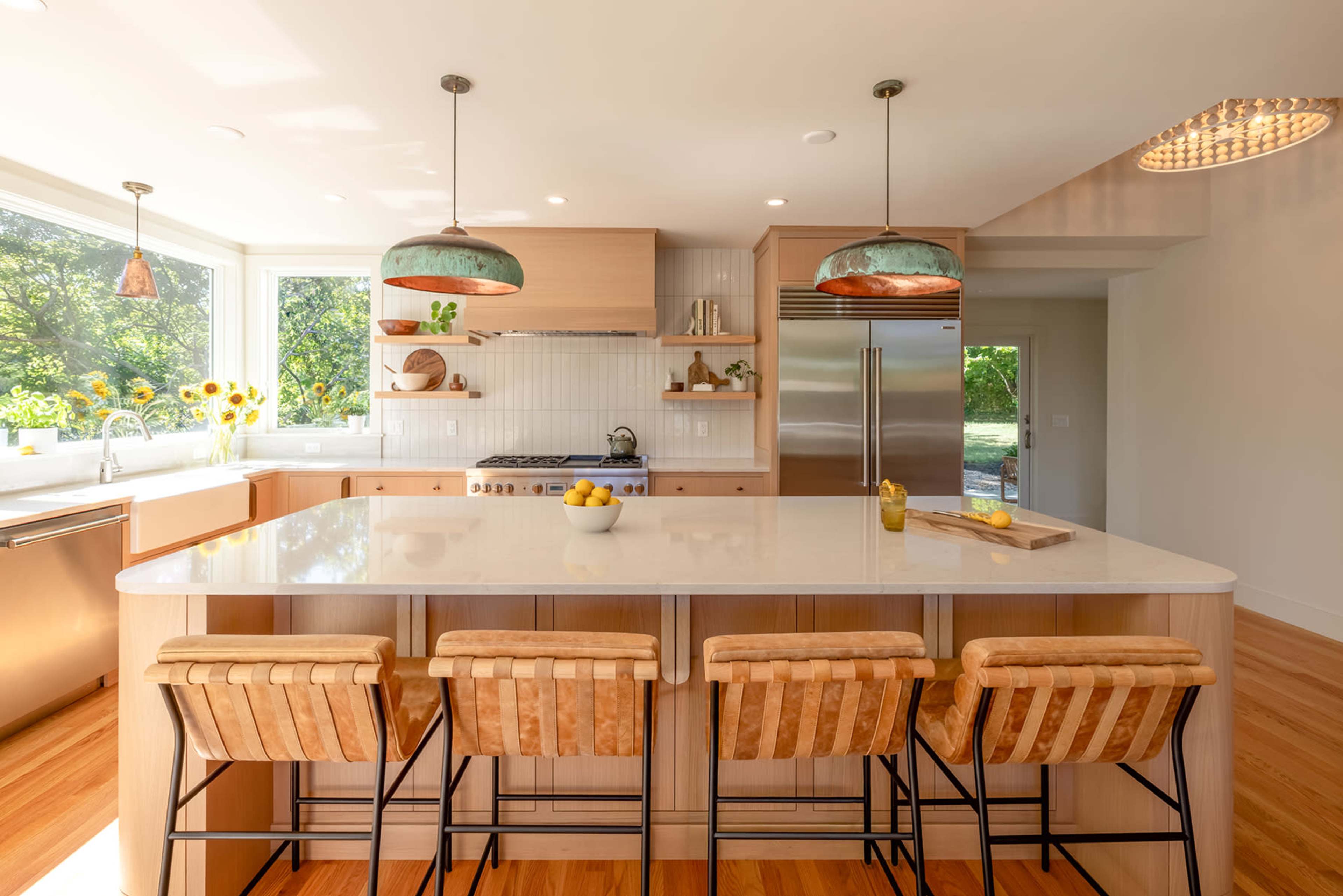 The image shows a modern kitchen with a large central island, three bar stools, and pendant lighting, complemented by a window view of greenery.