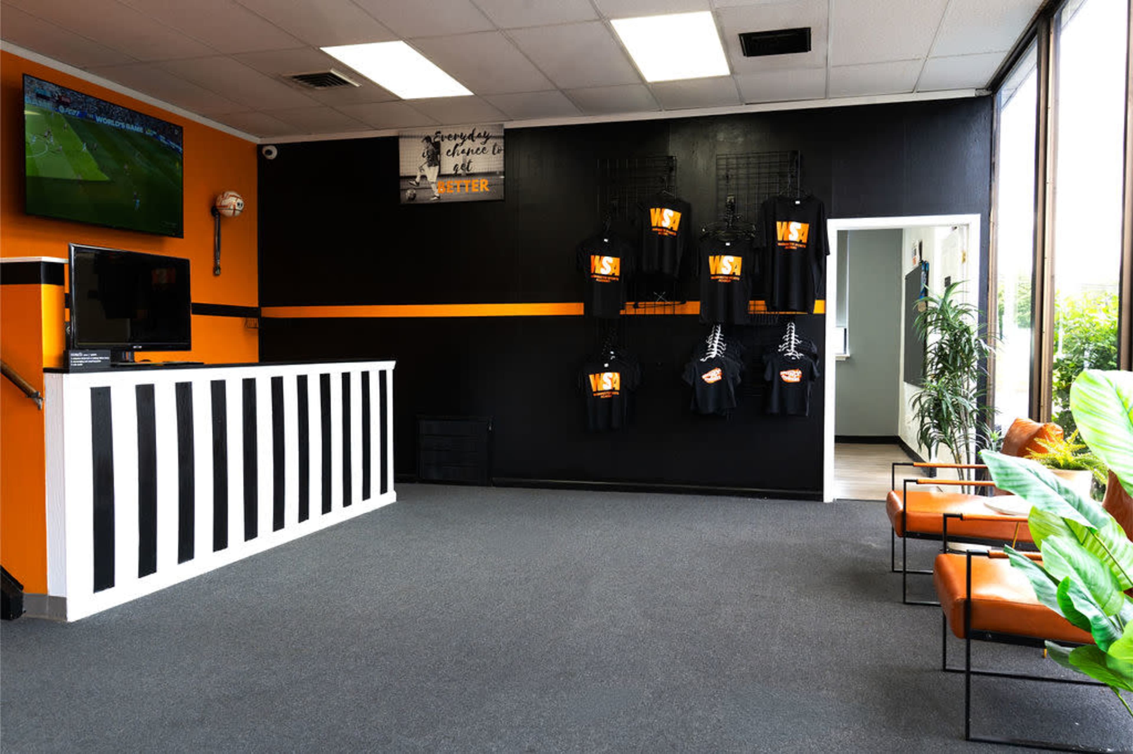 The image shows a spacious interior of a business with a black and orange color scheme, featuring a reception desk, a wall-mounted television, and display racks for merchandise.
