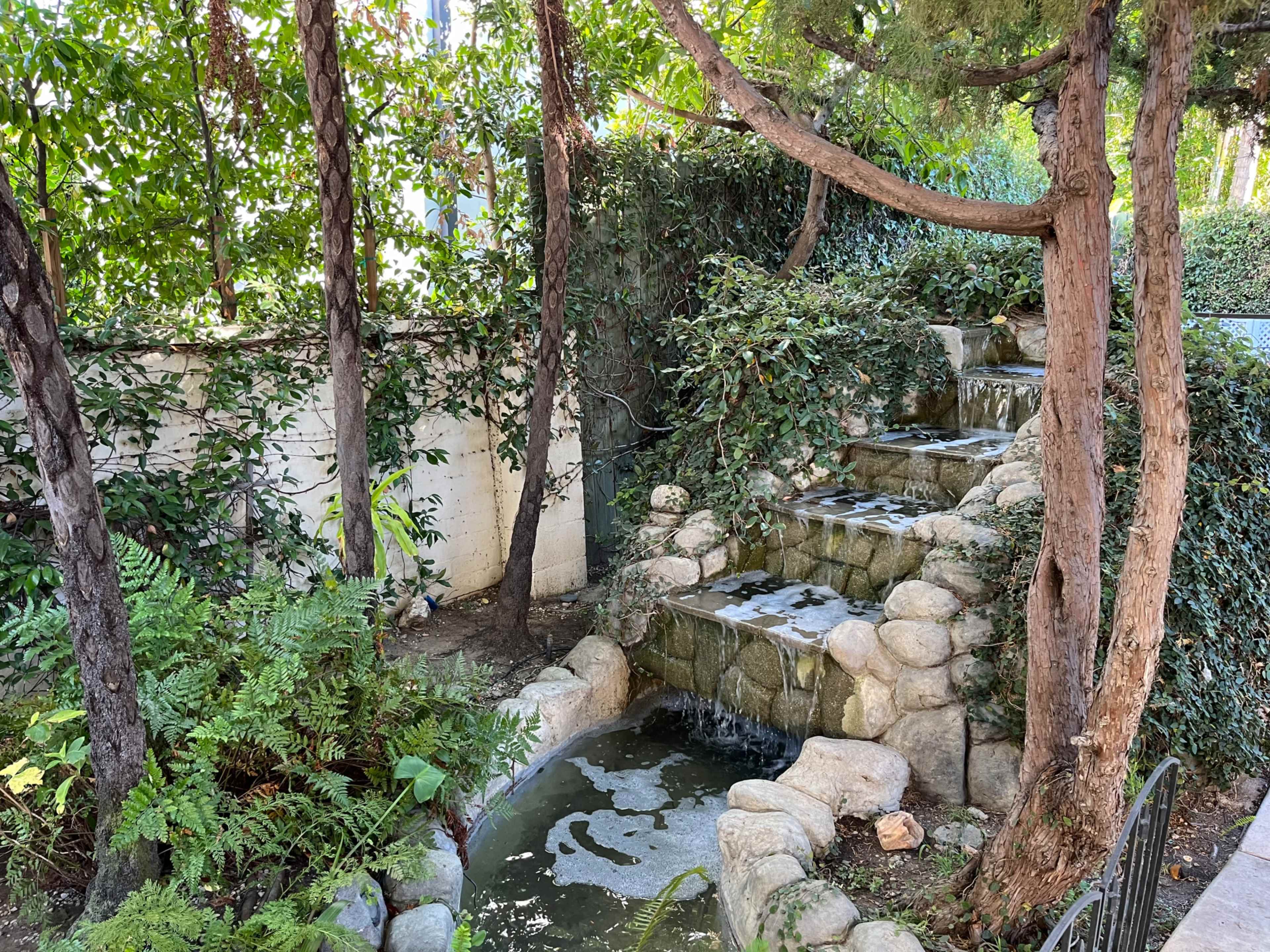 The image shows a serene landscaped area with a small stone waterfall flowing into a pond, surrounded by lush greenery and trees.