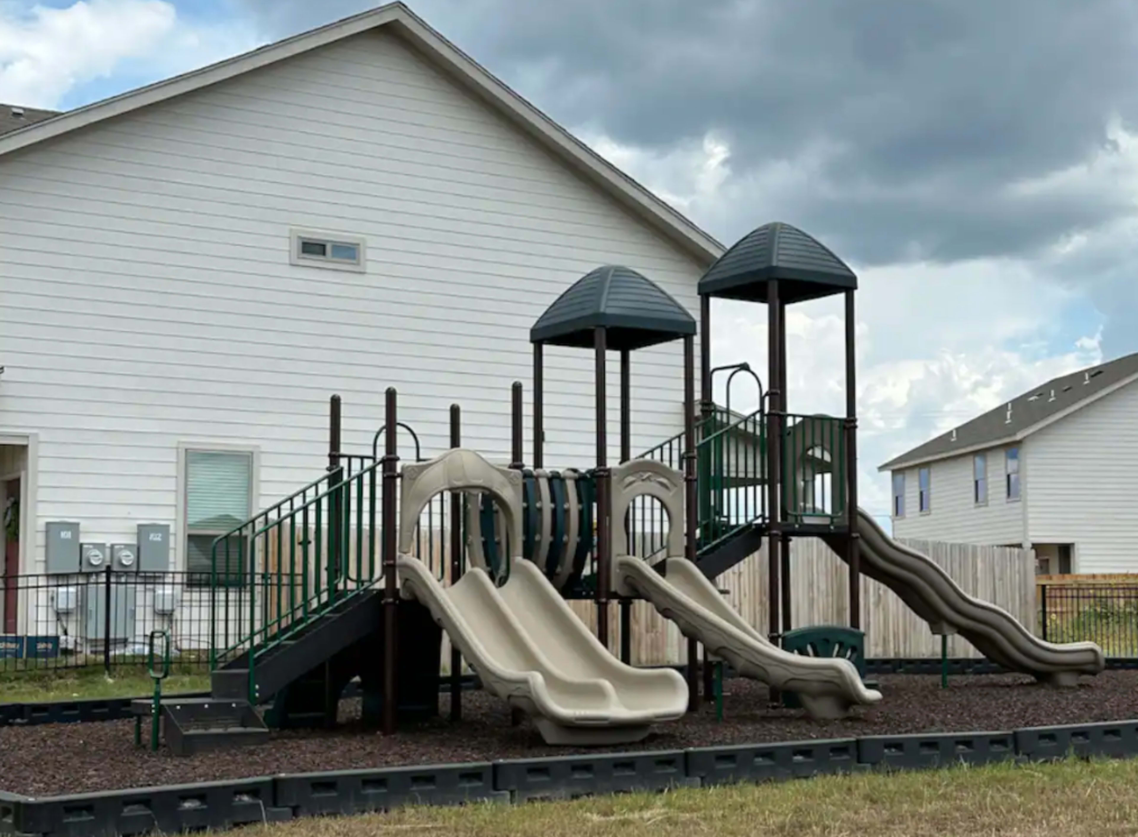 A playground with two slides and two play towers, situated next to a white house.