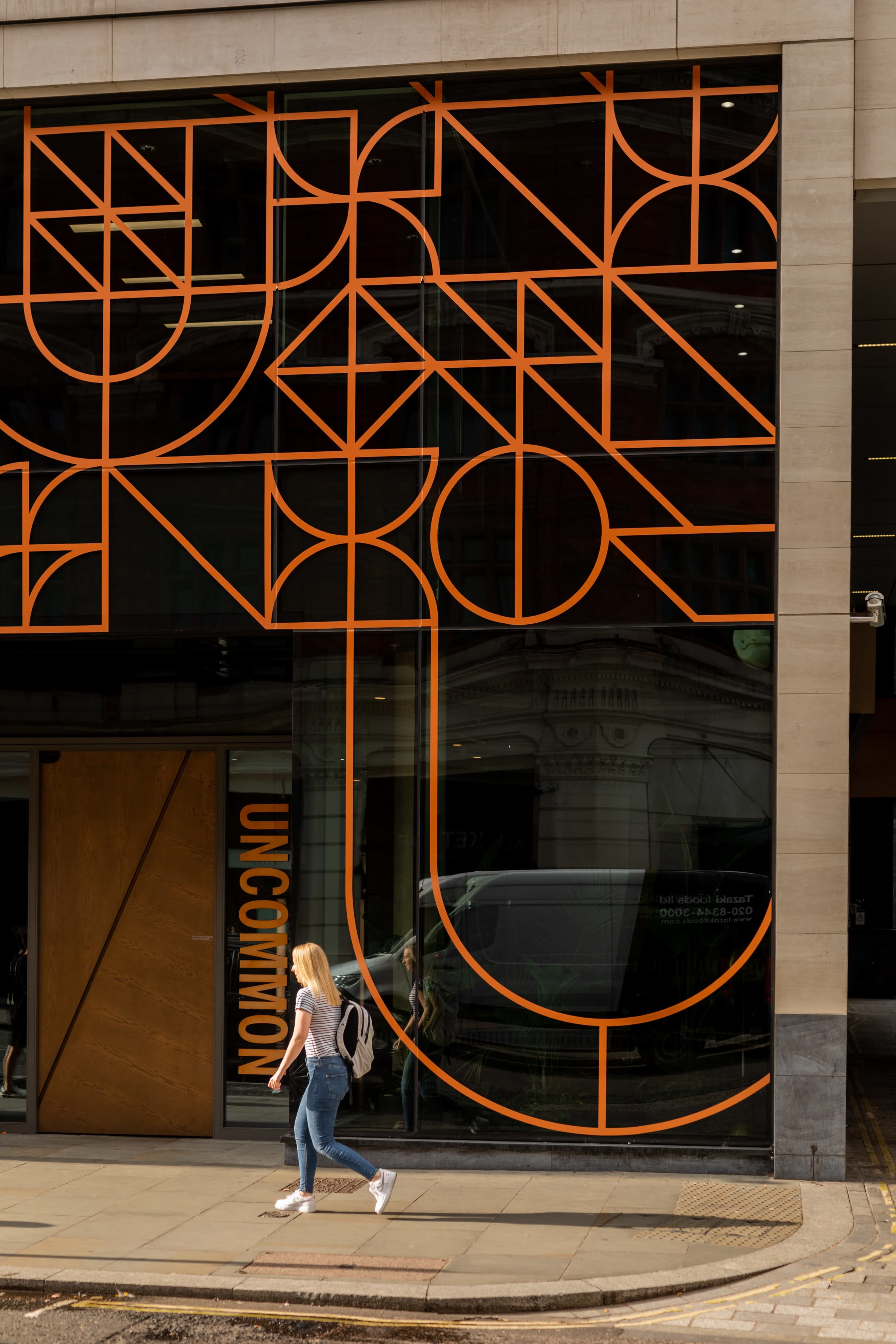 A woman with blonde hair walks past a building featuring large, orange geometric lettering on the glass facade.