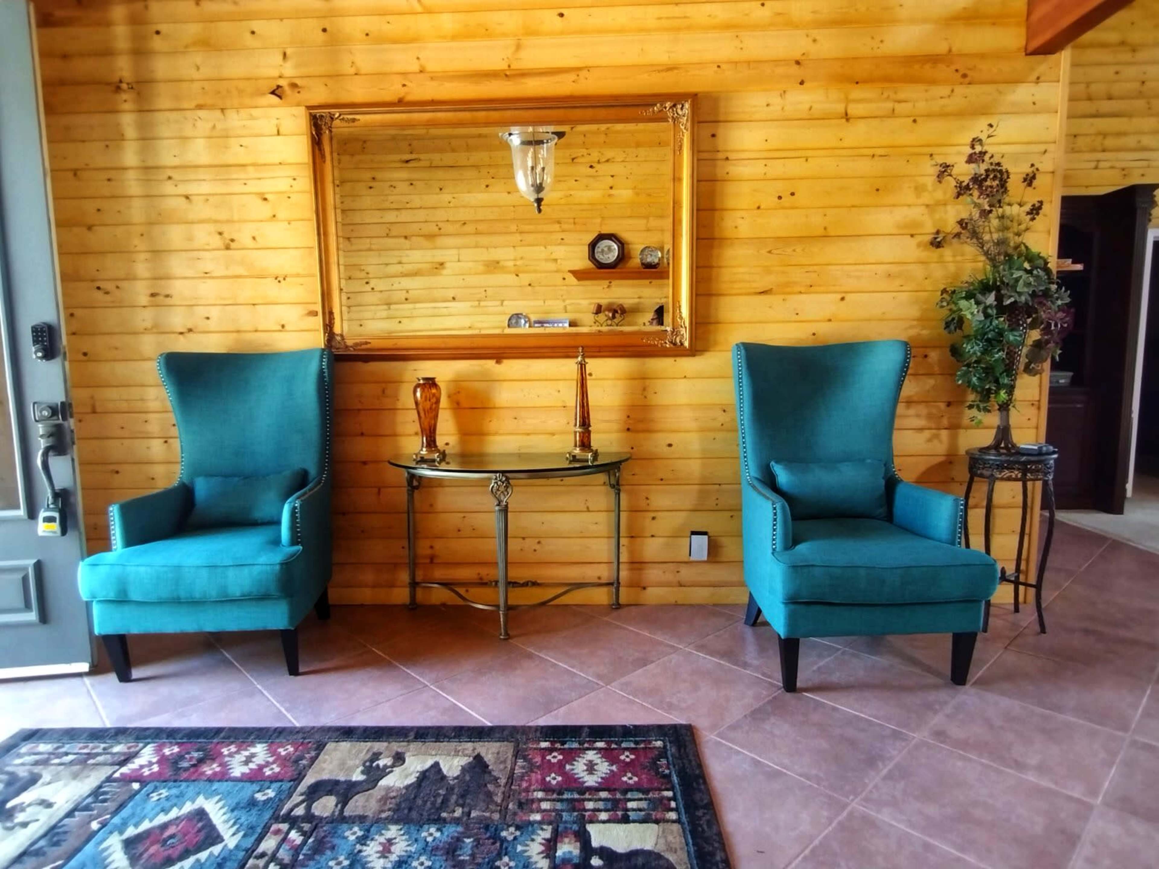 A cozy entryway featuring two teal armchairs, a circular glass table with decorative items, and a large mirror against wooden walls.