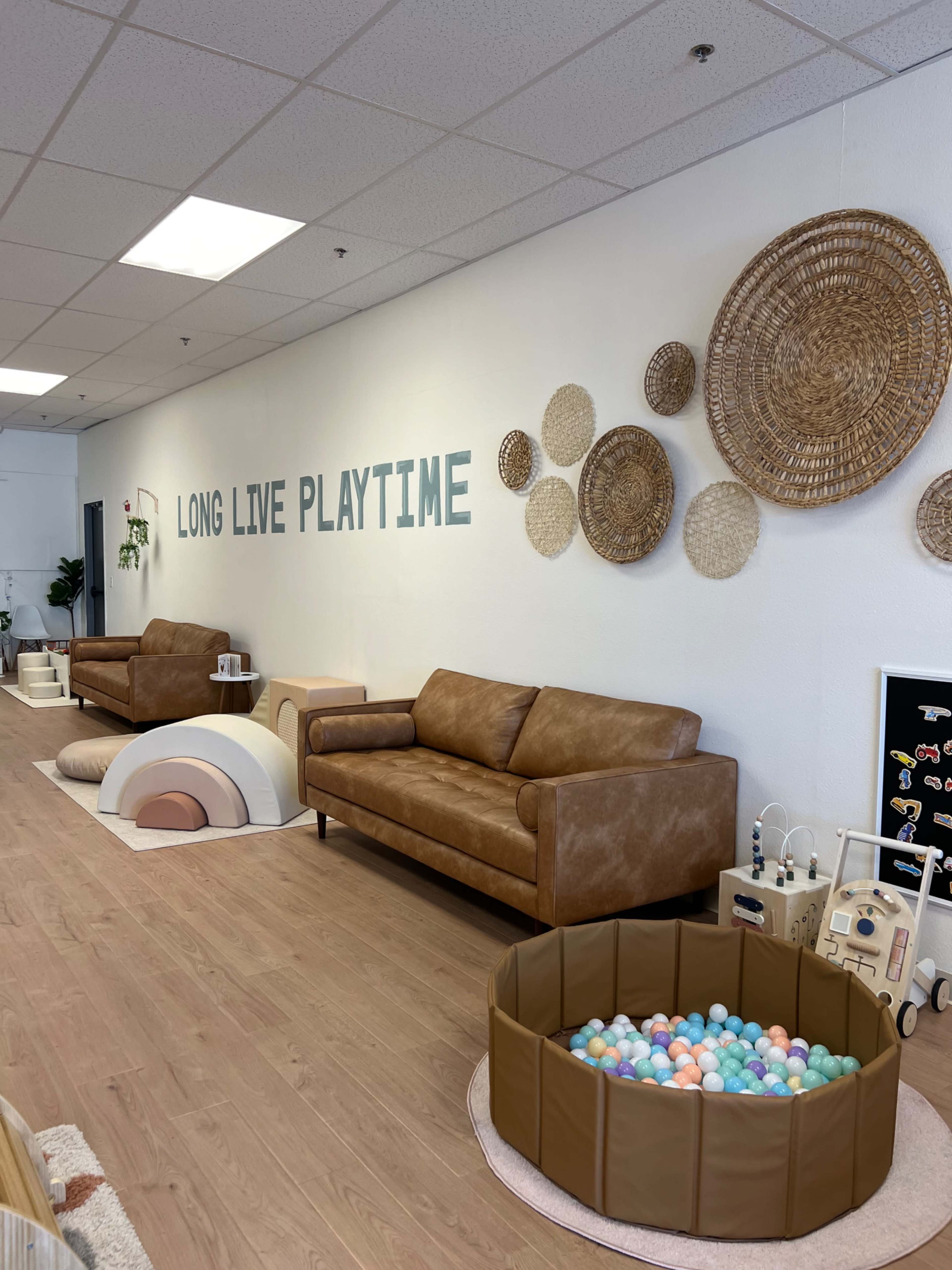 The image shows a brightly lit playroom with two brown leather sofas, decorative woven wall hangings, and a large ball pit filled with colorful balls.