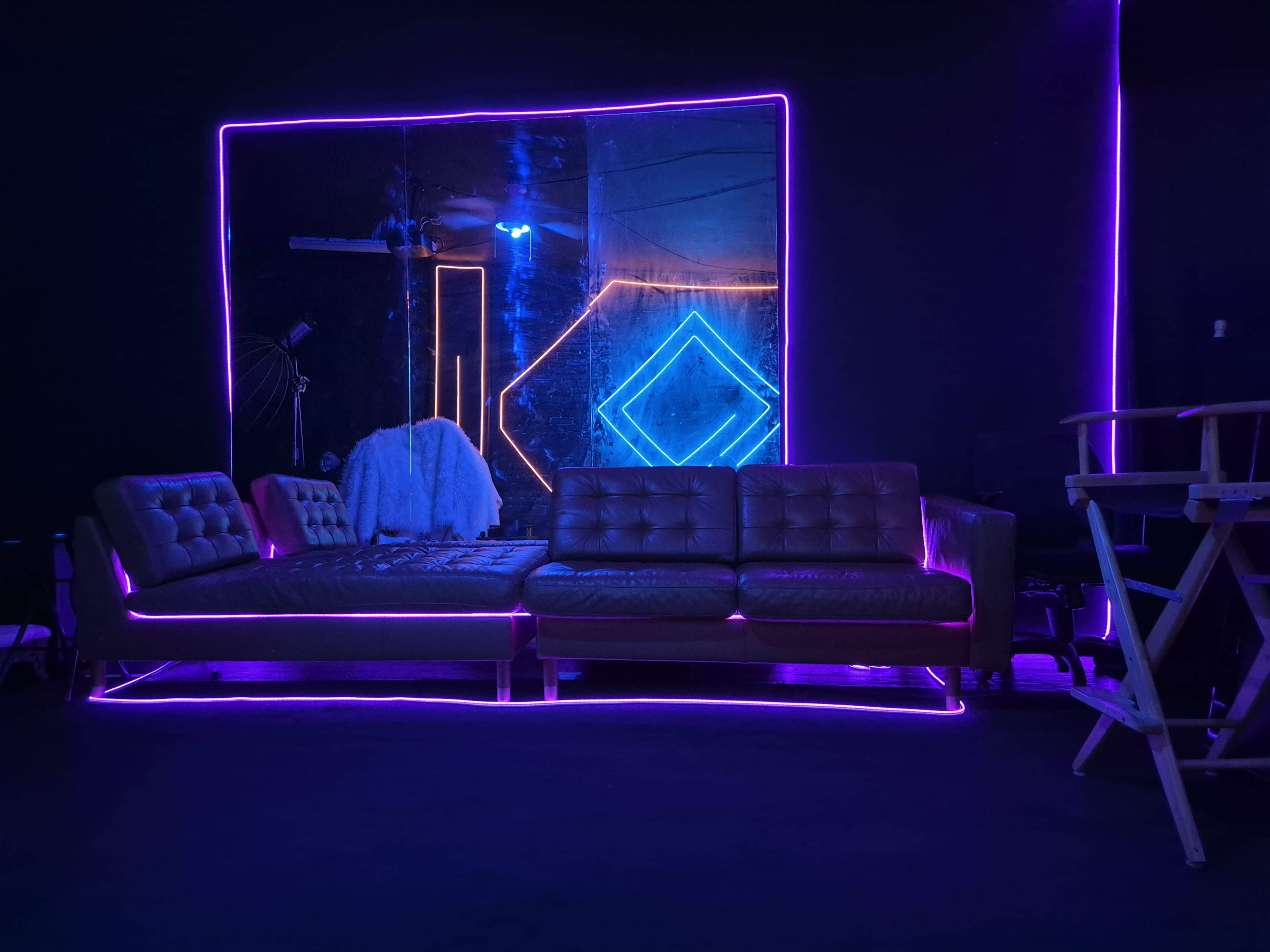 A modern lounge area features a brown leather sofa illuminated by neon pink and blue lights, with a large mirror reflecting the room's design elements.