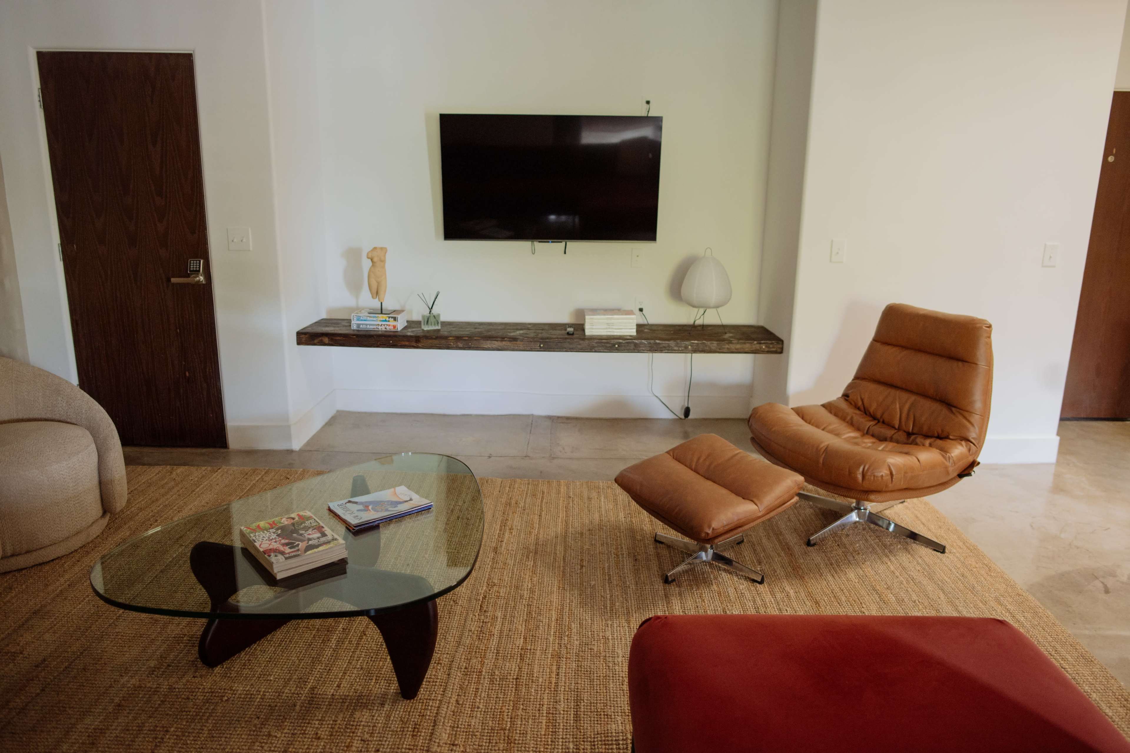 The living room features a glass coffee table, a brown lounge chair, a wooden shelf with decorative items, and a wall-mounted television.
