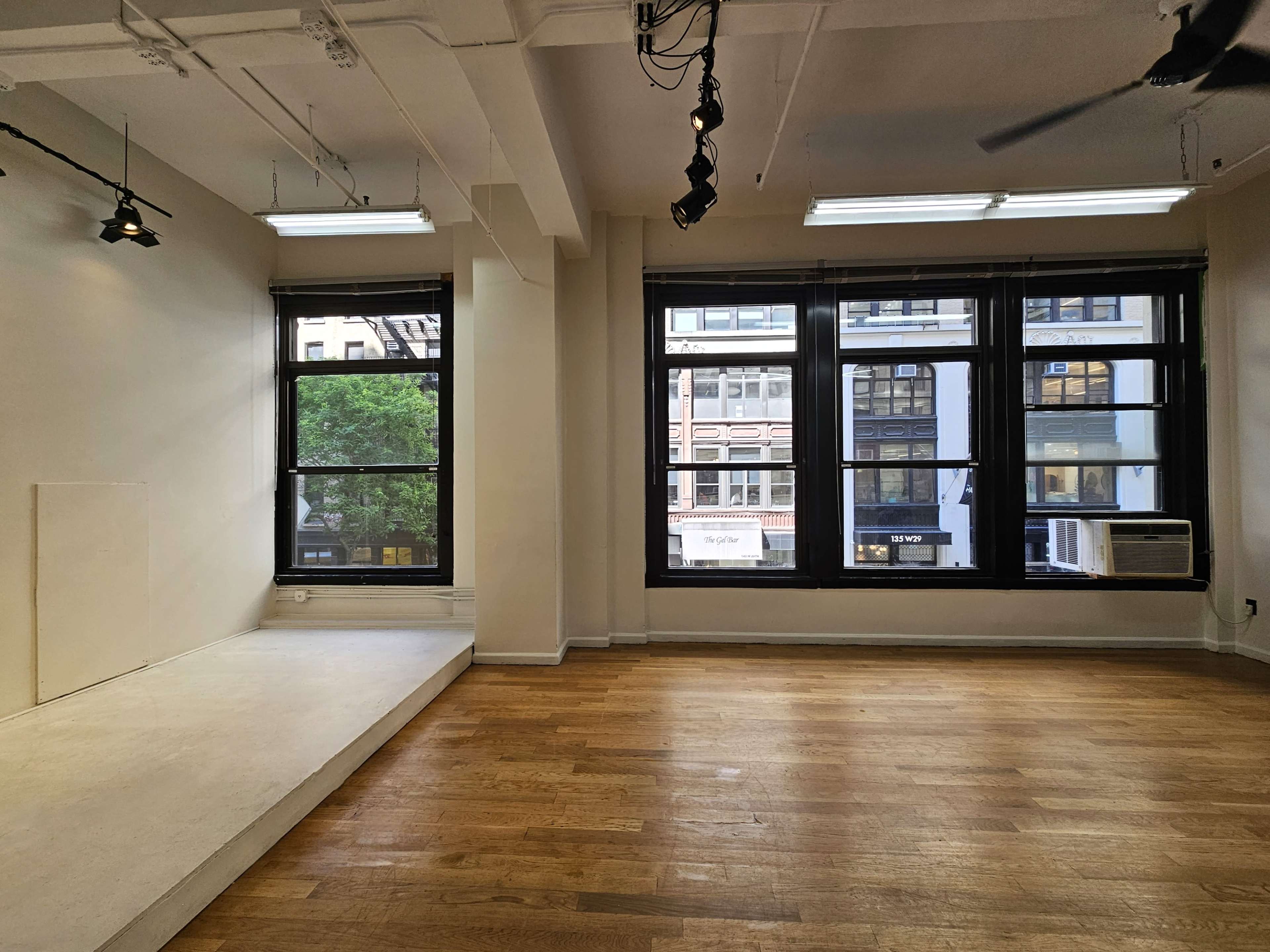 The image shows a spacious empty room with wooden flooring, large windows revealing a view of buildings outside, and a small elevated area on one side.