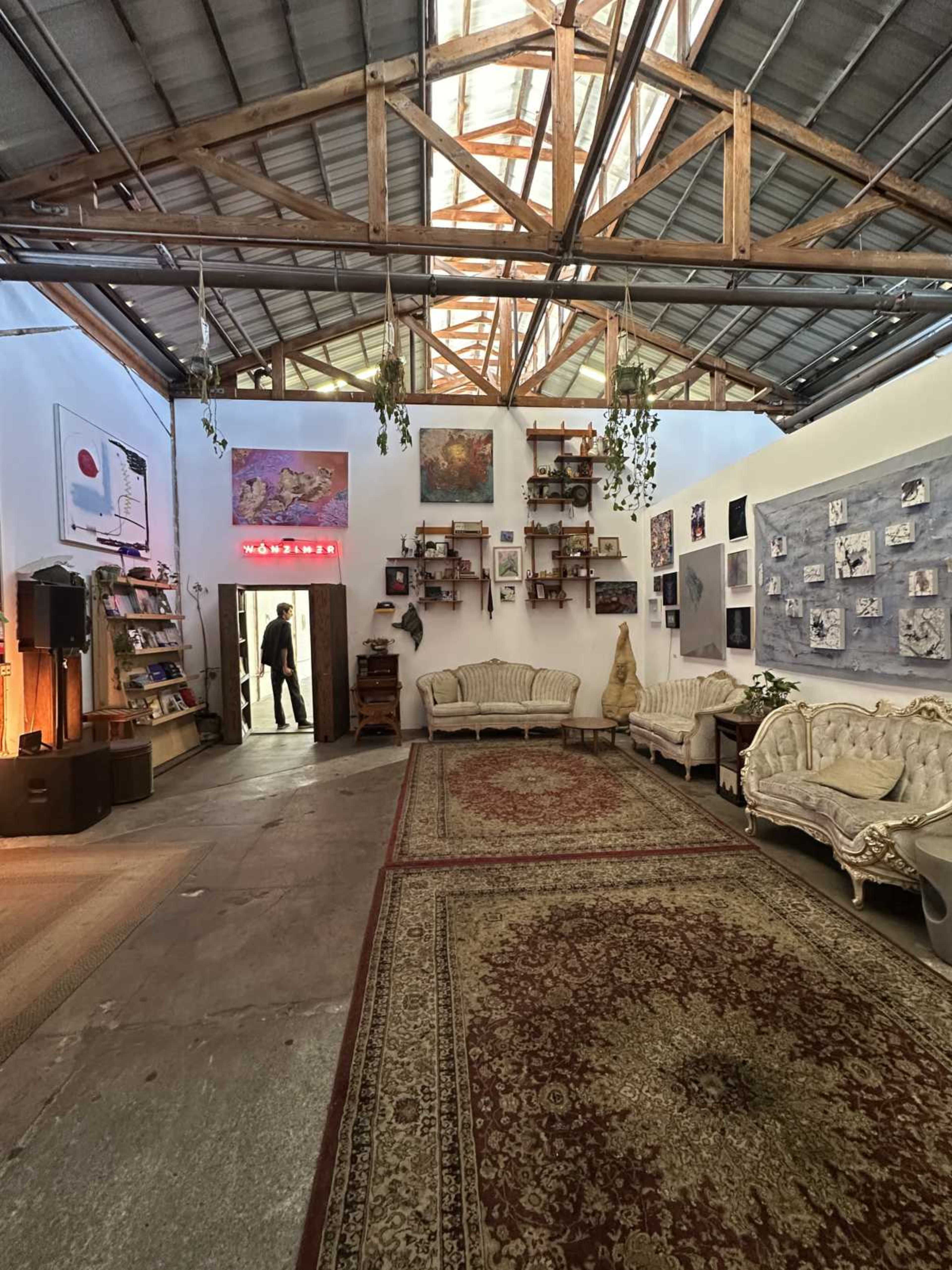 The image features a spacious art studio or gallery with vintage furniture, a patterned rug, and shelves adorned with pictures and plants, set under a skylit ceiling.