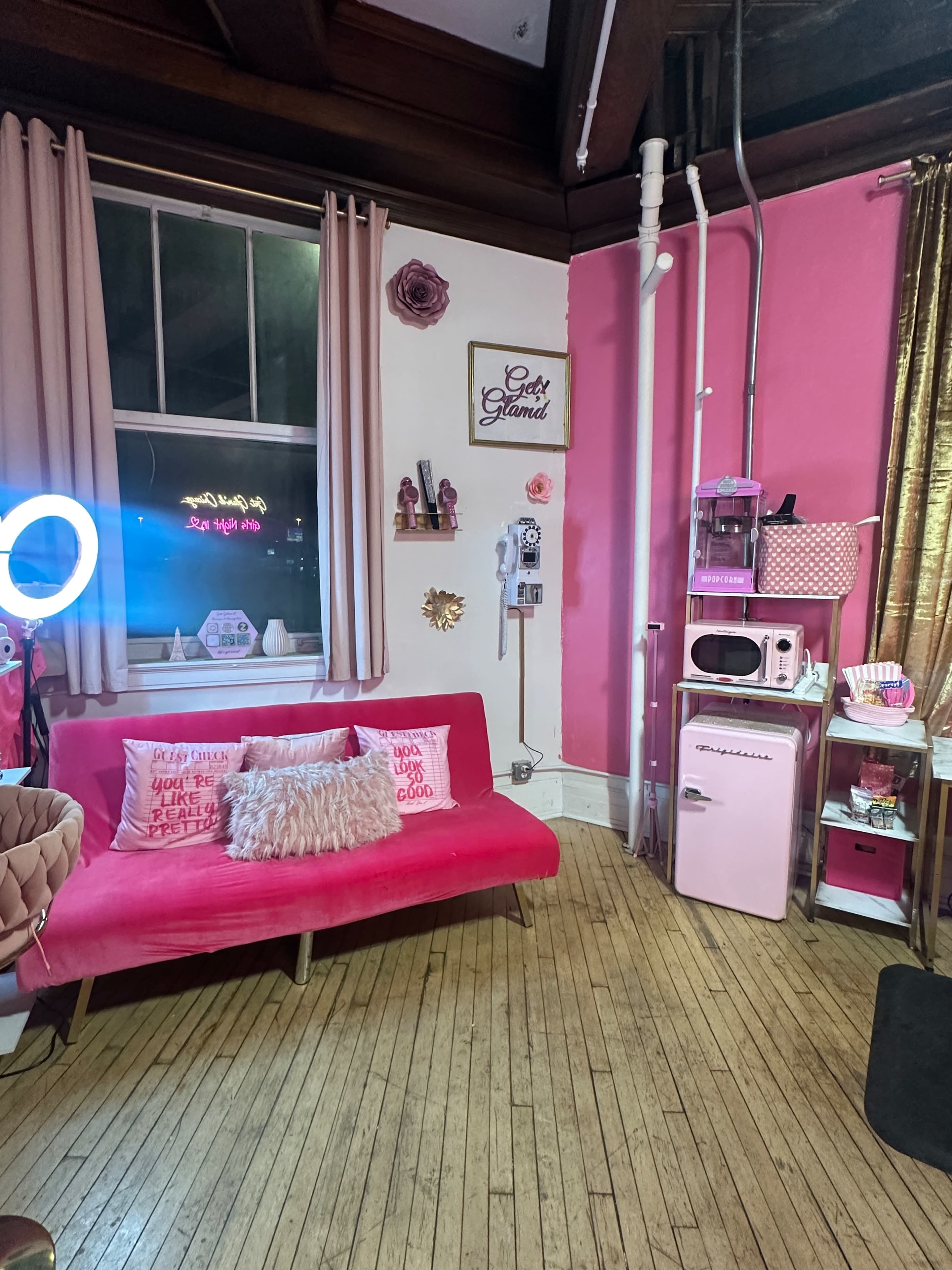 The image depicts a vibrant pink-themed room featuring a pink couch, curtains, and wall decorations, along with a kitchenette area that includes a retro-style microwave and refrigerator.