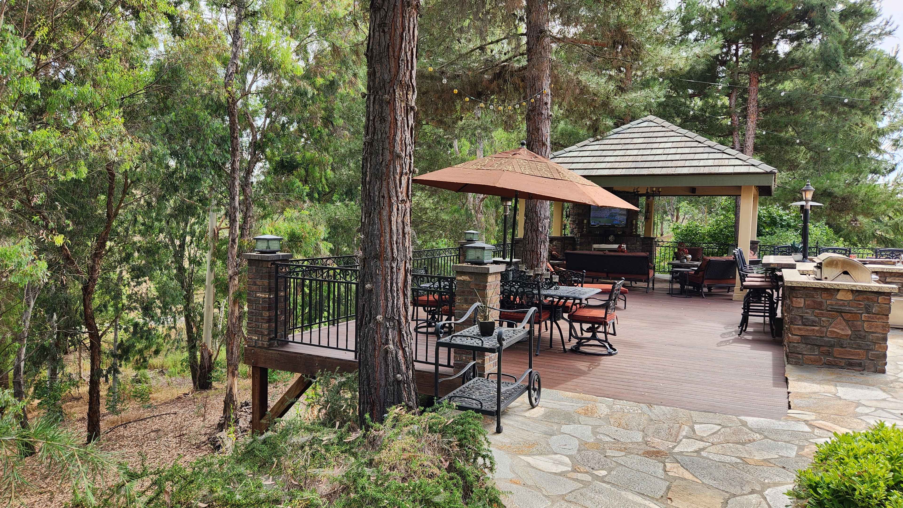 A wooden deck with metal furniture and umbrellas overlooks a forested area, adjacent to a covered seating area.