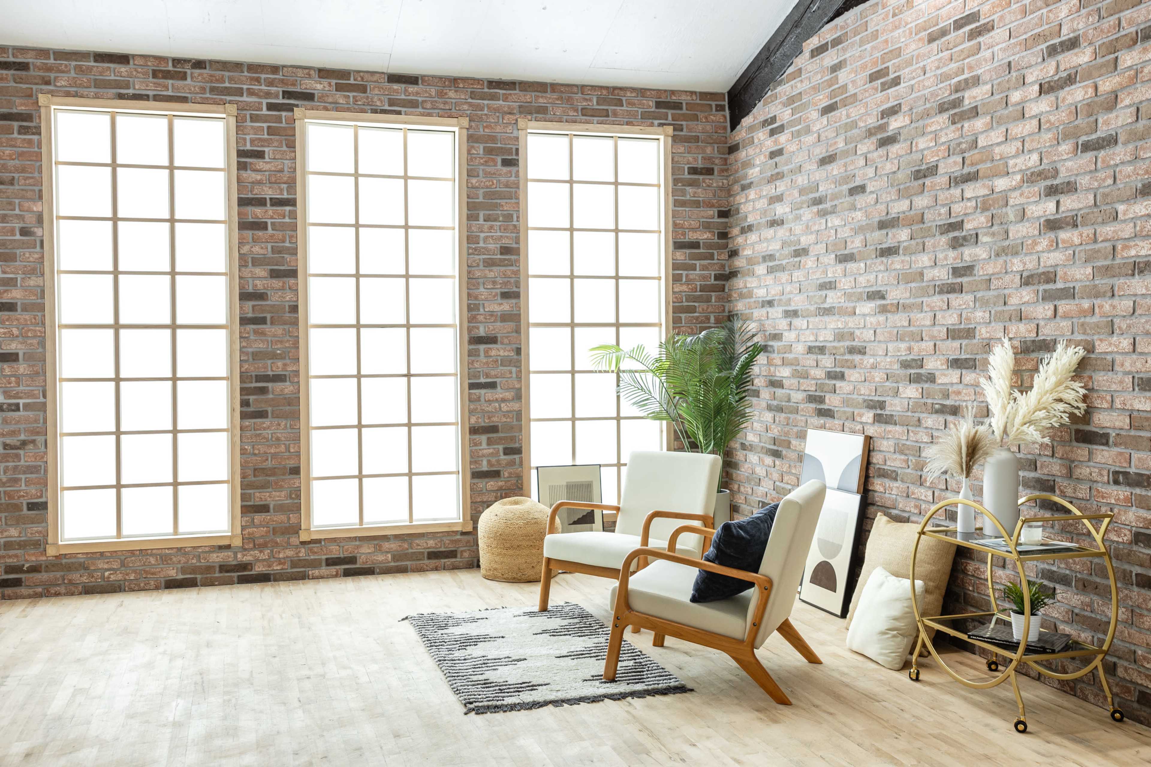 A modern interior space features large windows, a brick wall, a pair of armchairs, a decorative plant, and a gold cart.