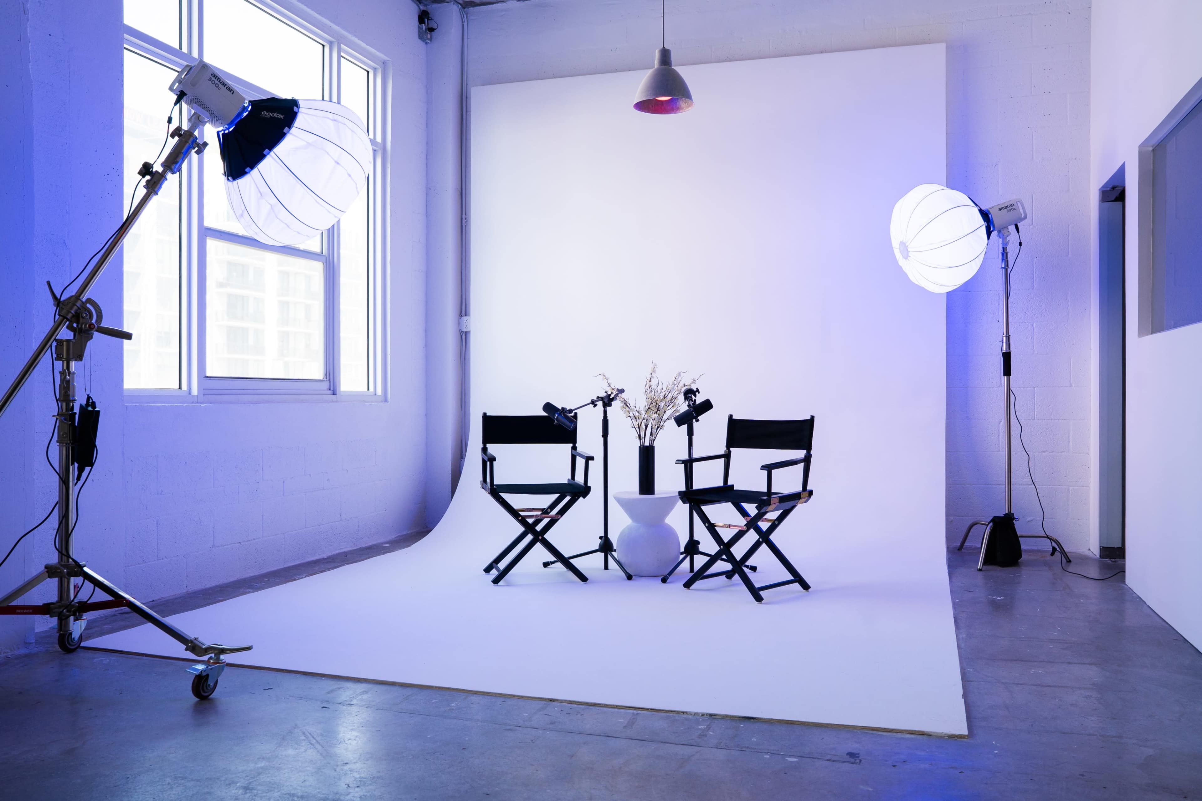 A photography studio features two director's chairs positioned on a white backdrop, with studio lights and a vase of dried flowers in the center.