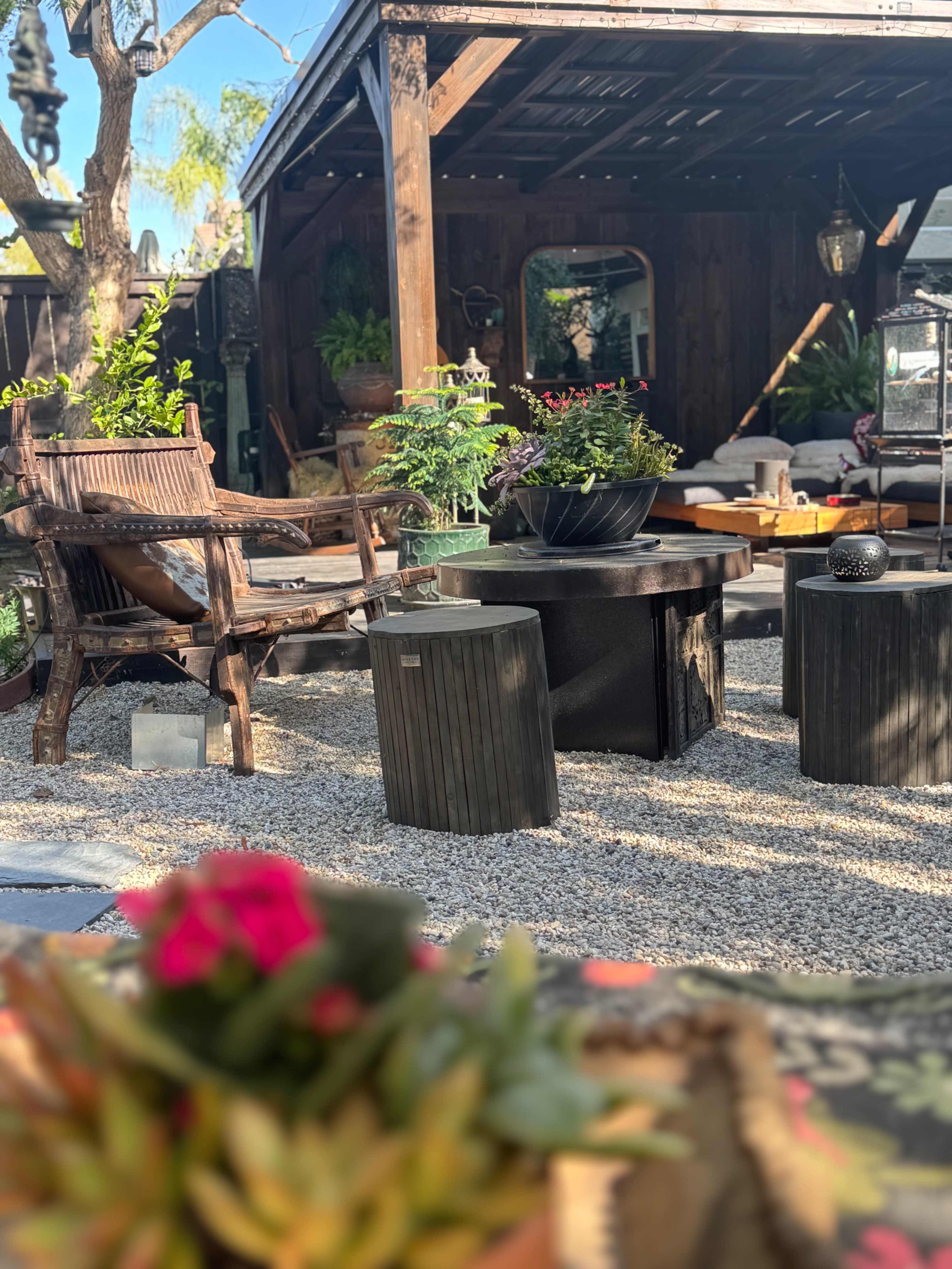 The image shows a cozy outdoor seating area with wooden furniture, potted plants, and gravel flooring.