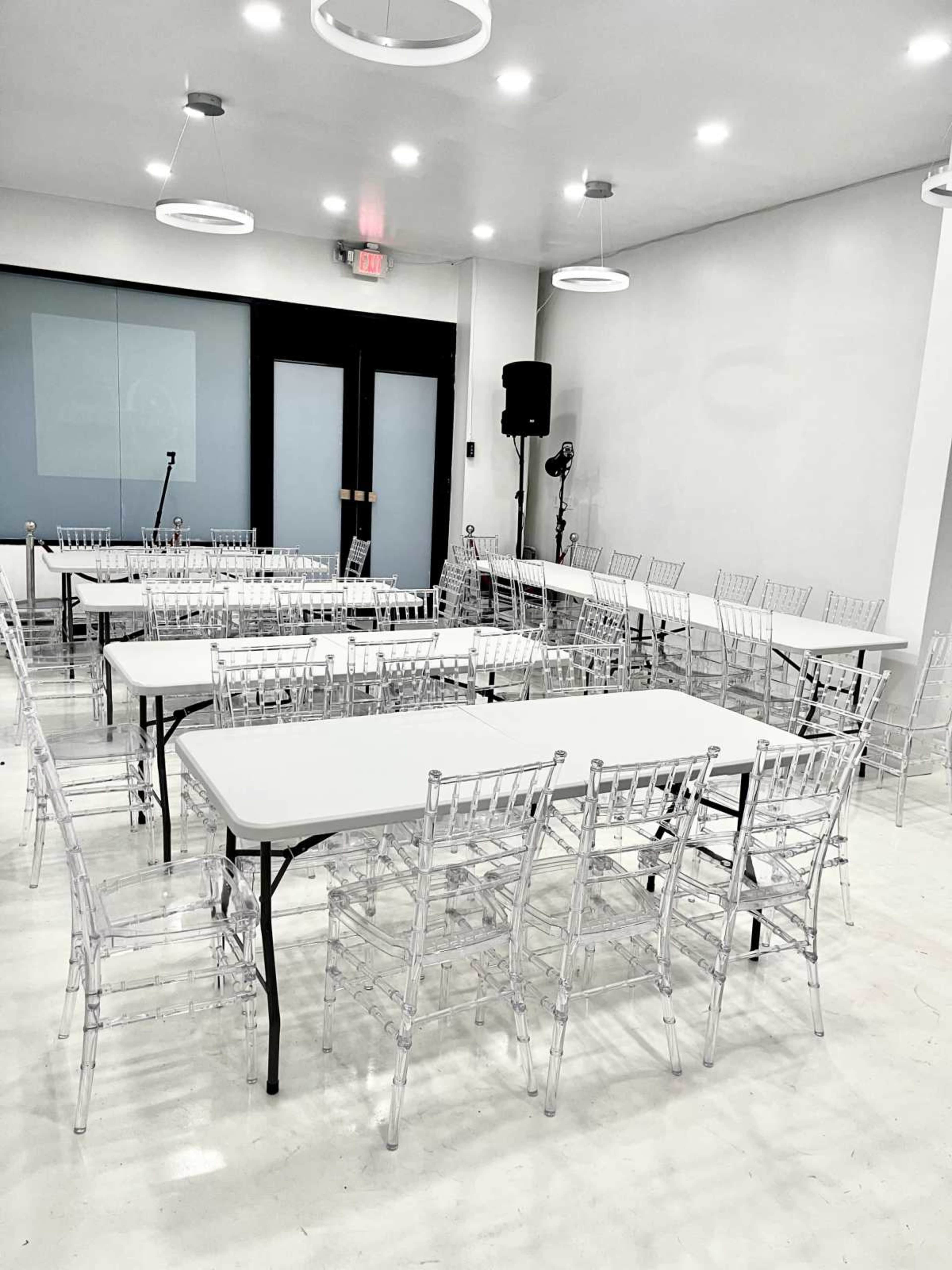 A spacious room is set up with several rectangular tables and clear acrylic chairs arranged in a circle, featuring a microphone and speaker in the corner.