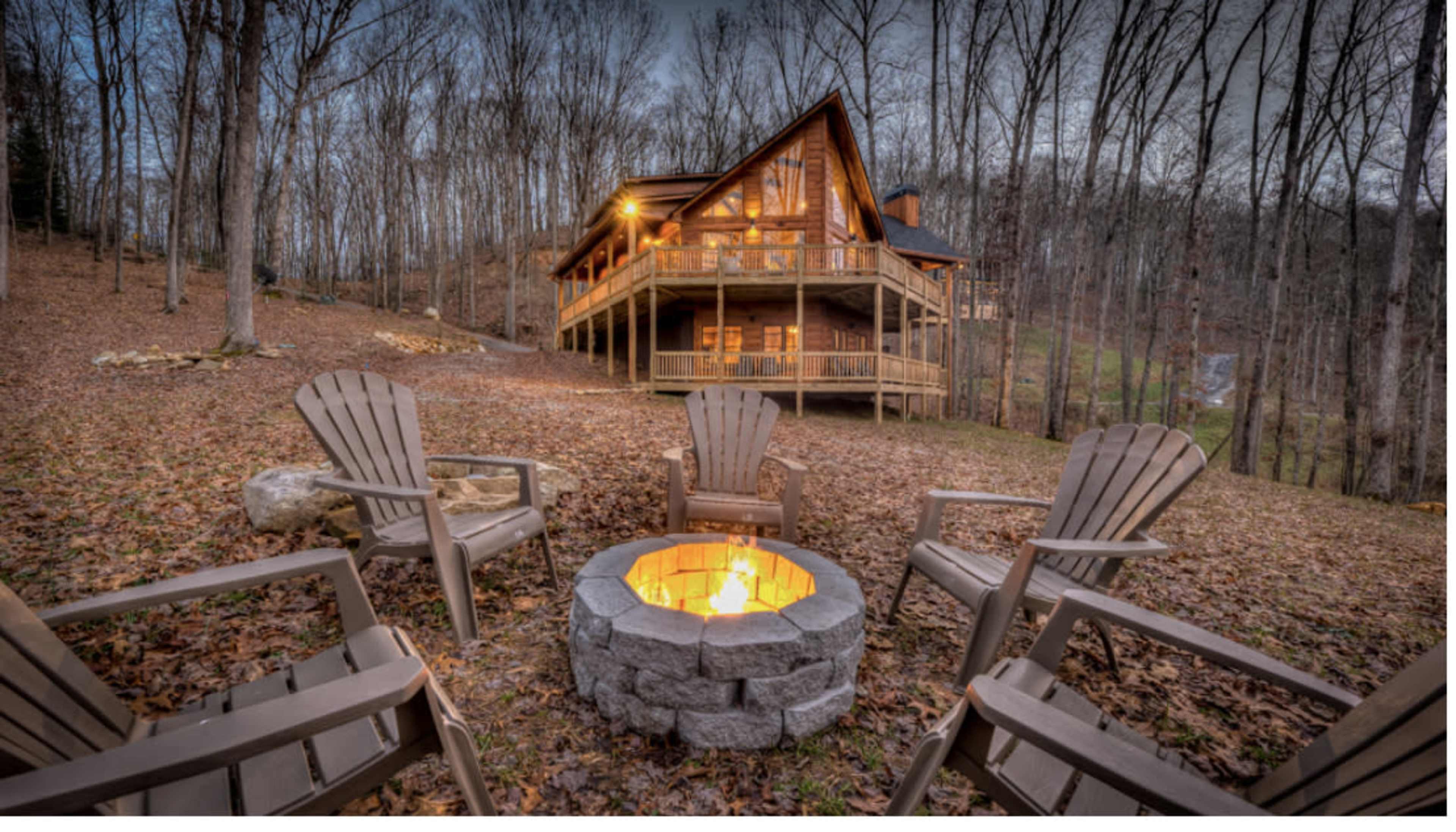 A cozy cabin surrounded by trees features a firepit with four chairs arranged around it.