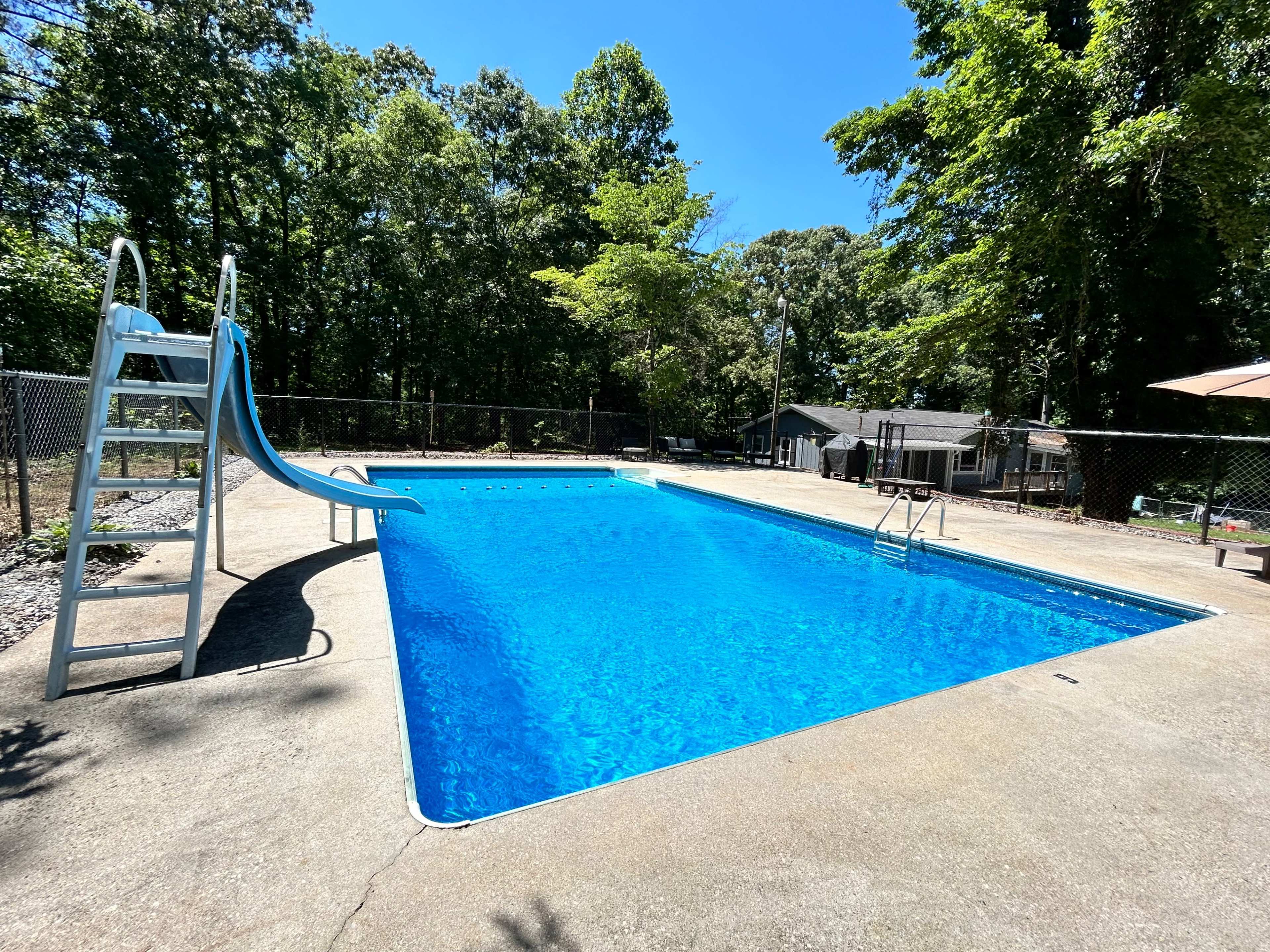 A clear blue swimming pool with a slide and diving board is surrounded by trees and a fence.