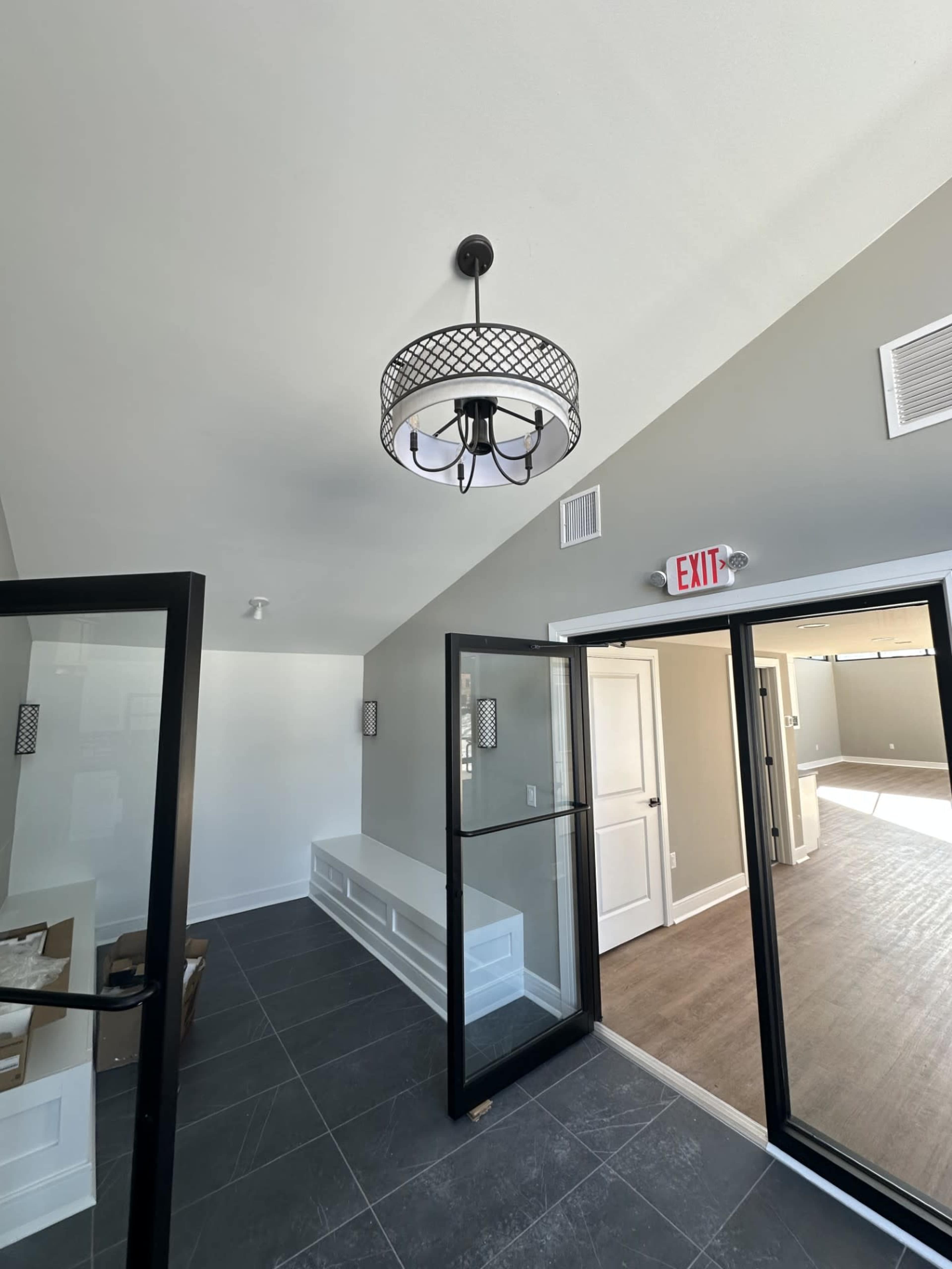 The image shows an entryway with a modern chandelier, gray walls, and black-framed glass doors leading to a room with wood flooring.