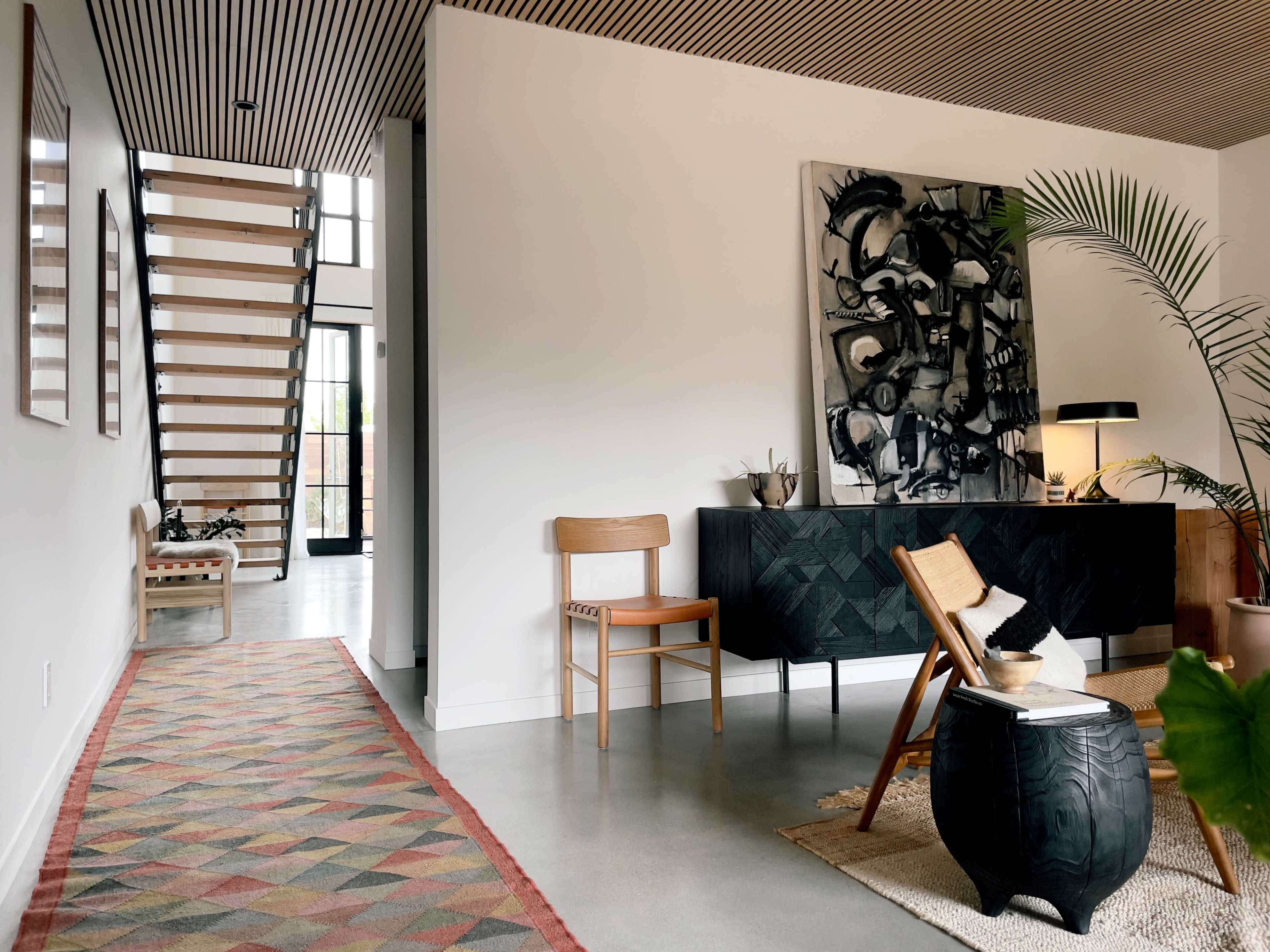 A modern interior hallway featuring a staircase, an artwork on the wall, a chair, a textured cabinet, and a patterned rug.