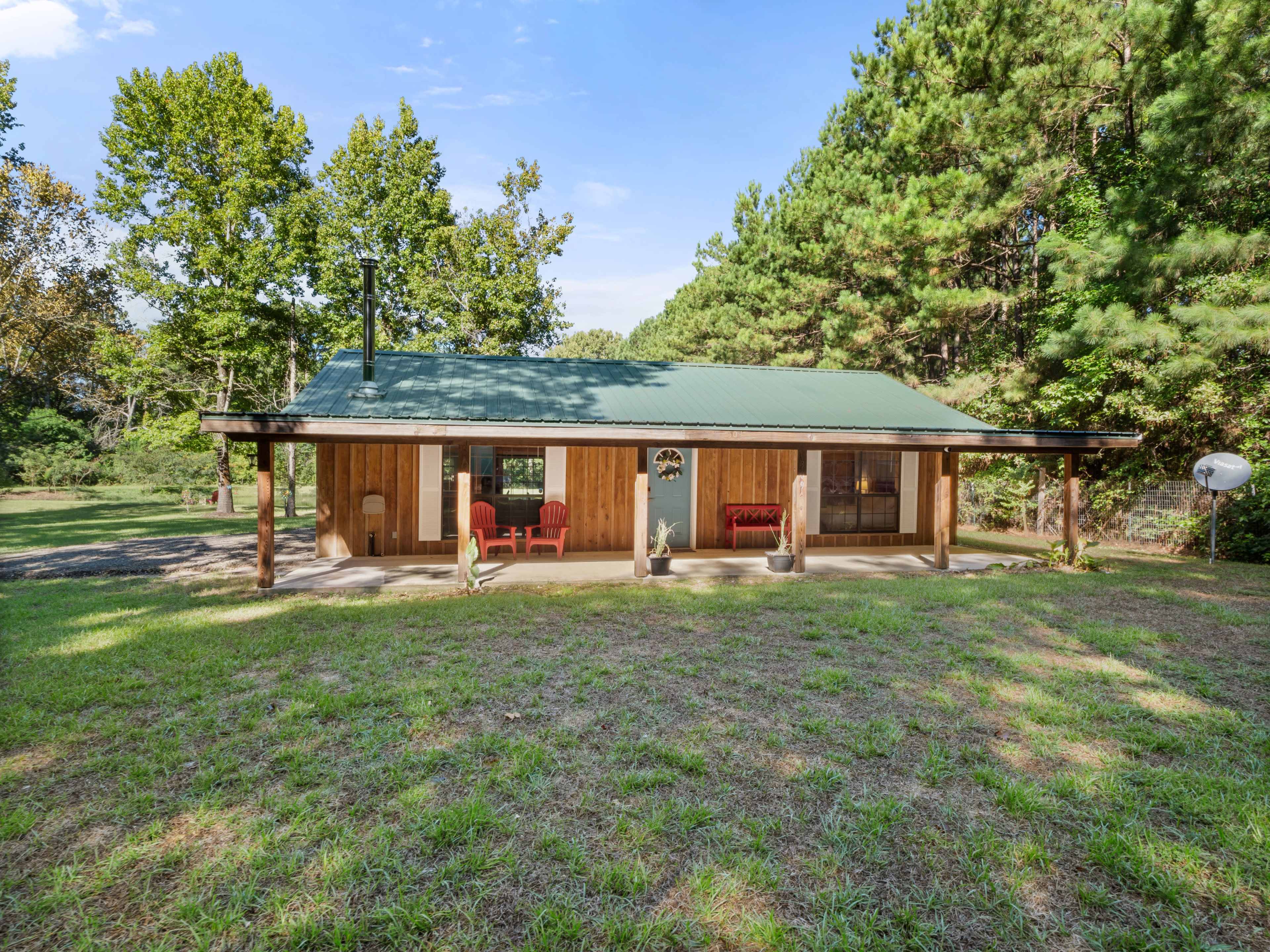 A single-story wooden cabin with a green metal roof and a porch, surrounded by trees and open grass.