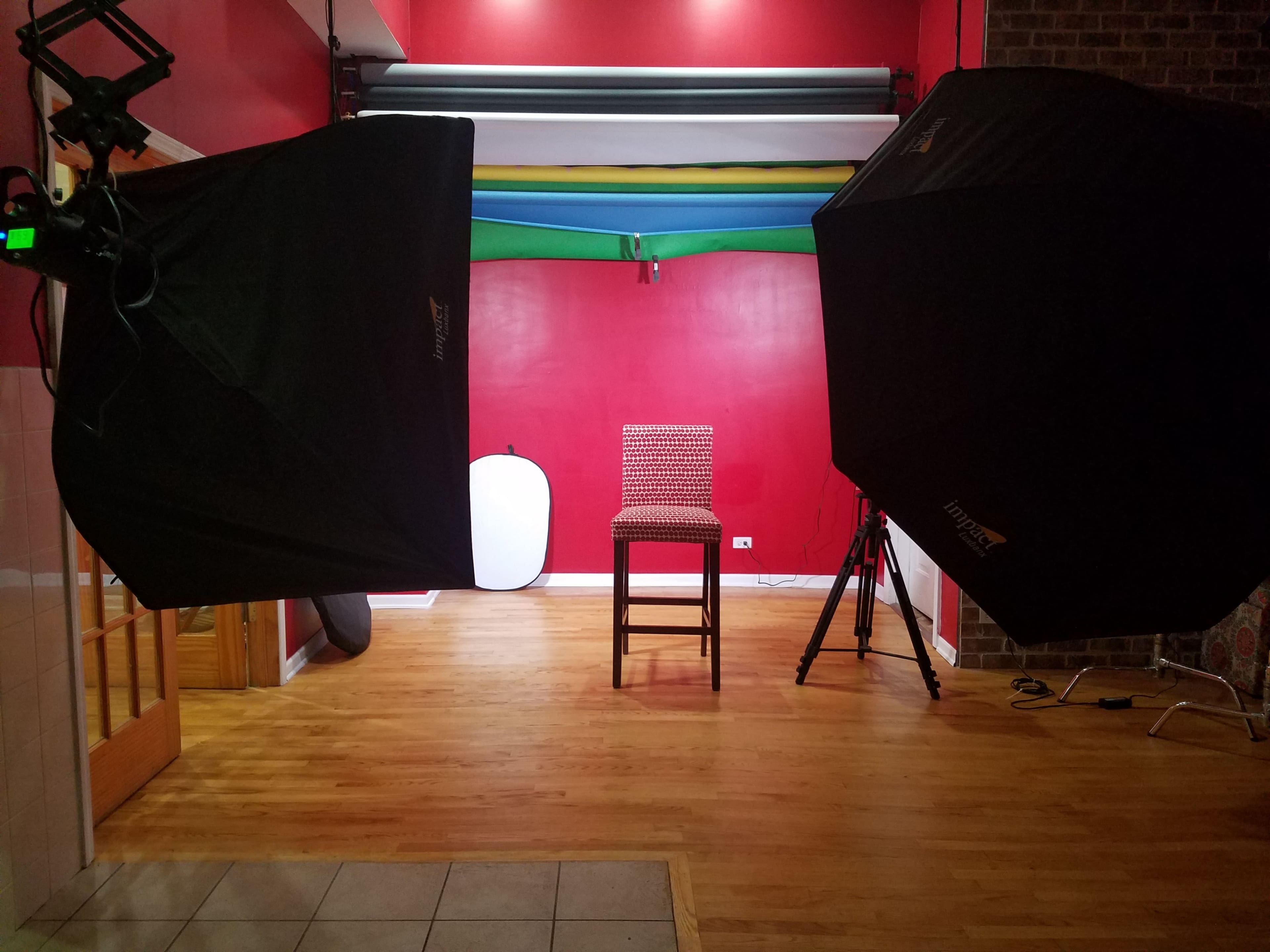 A photography studio features a single red chair set between two large softbox lights on either side, with colorful backdrops hanging above.