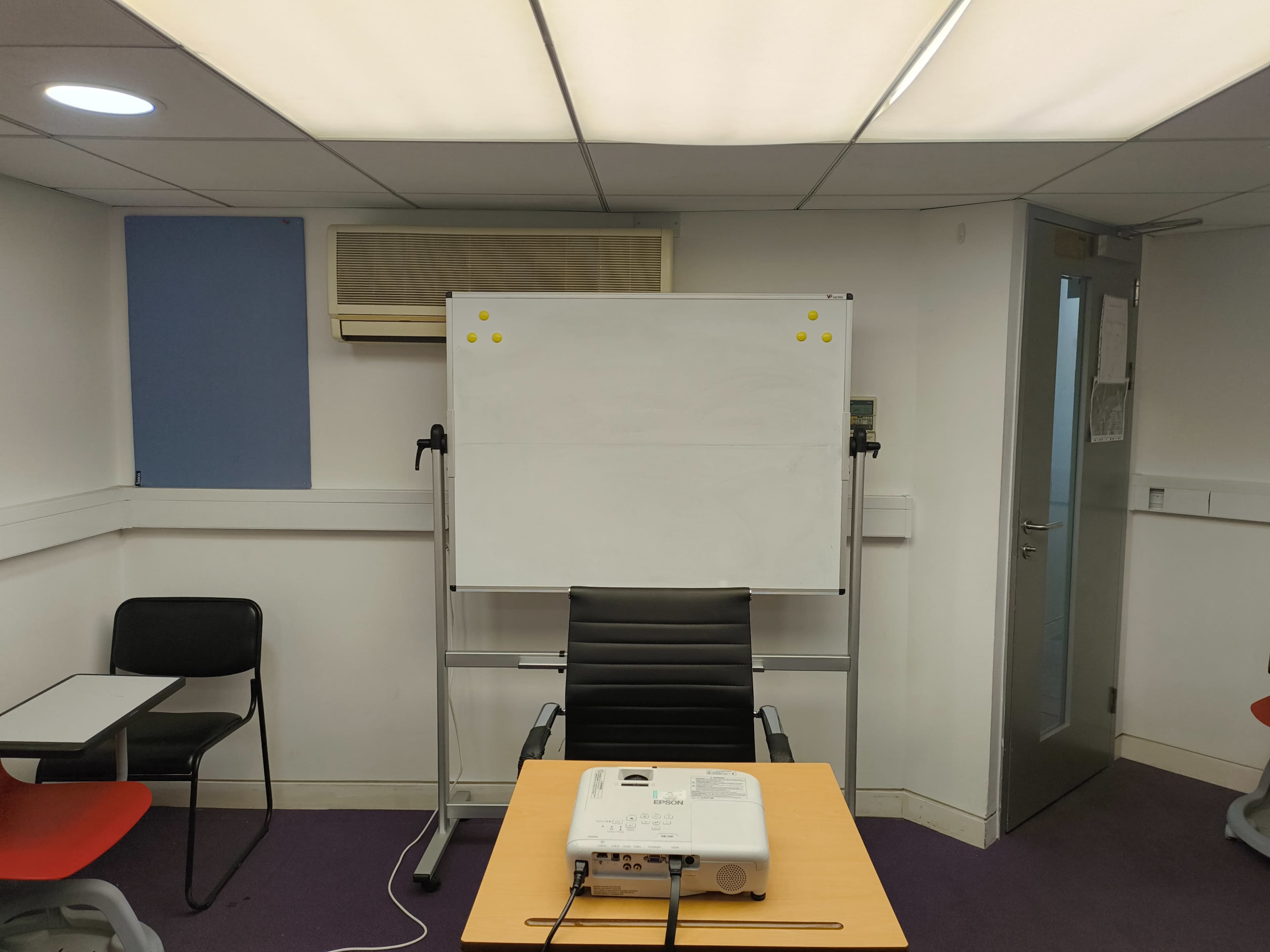 The image shows a small meeting room with a whiteboard on a stand, a projector on a table, and a black office chair facing the board.