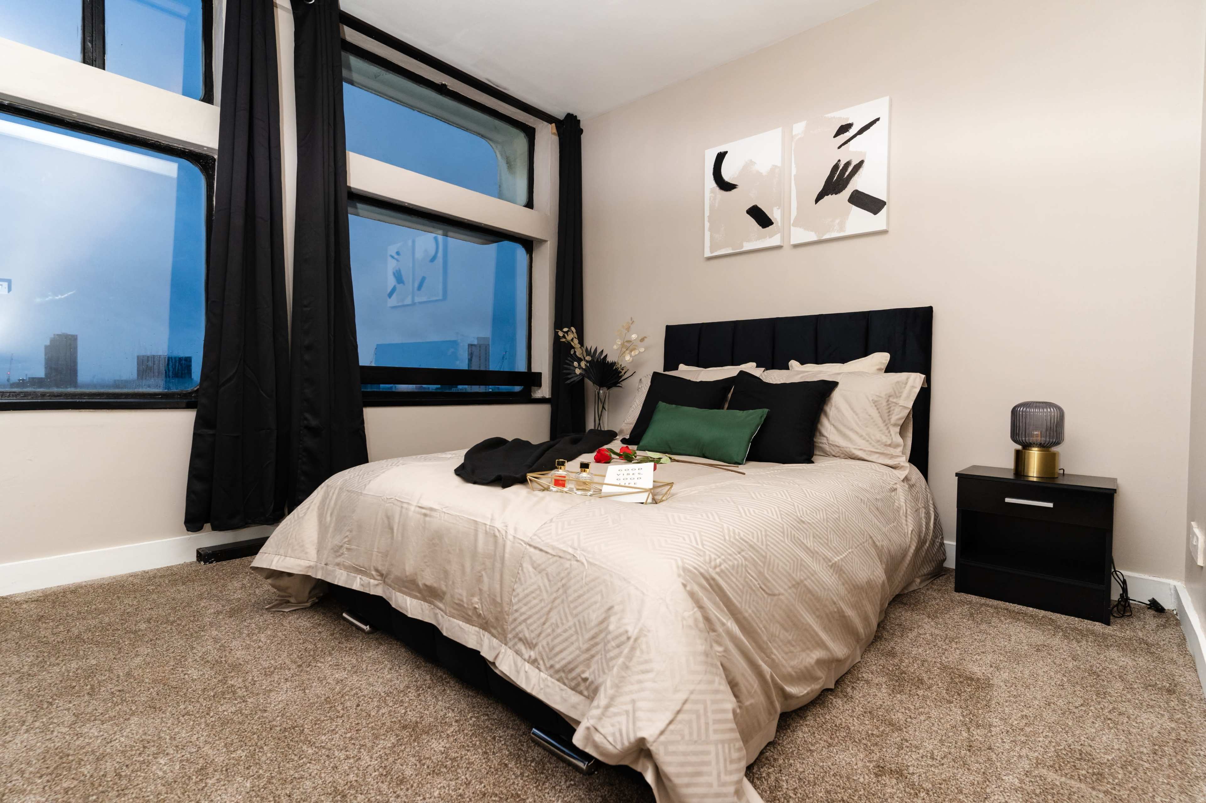 A neatly arranged bedroom features a large bed with a beige quilt, a nightstand with a lamp, and large windows dressed with black curtains.