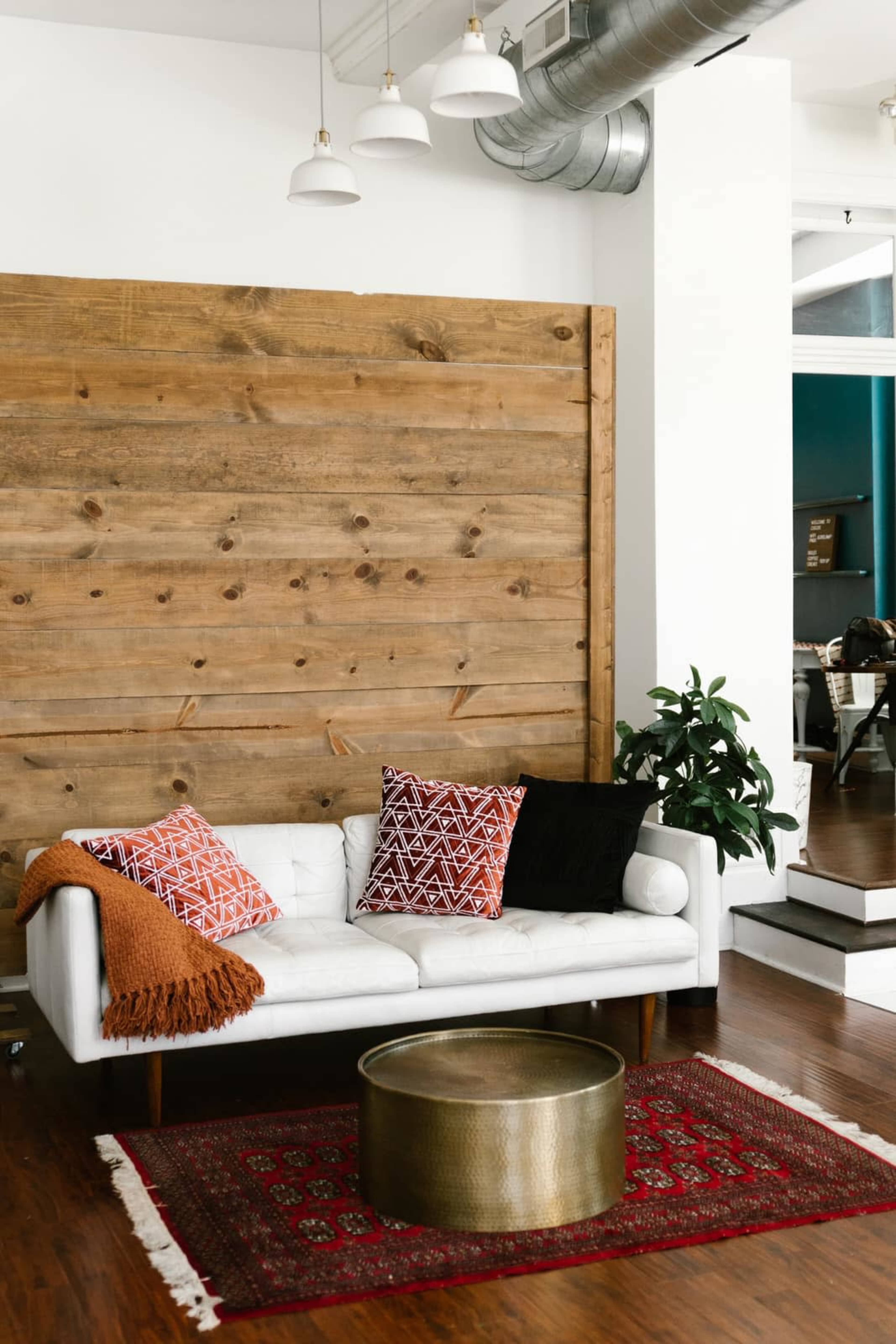 The image shows a modern living room with a white couch adorned with patterned pillows, a brass coffee table, and a wooden accent wall.