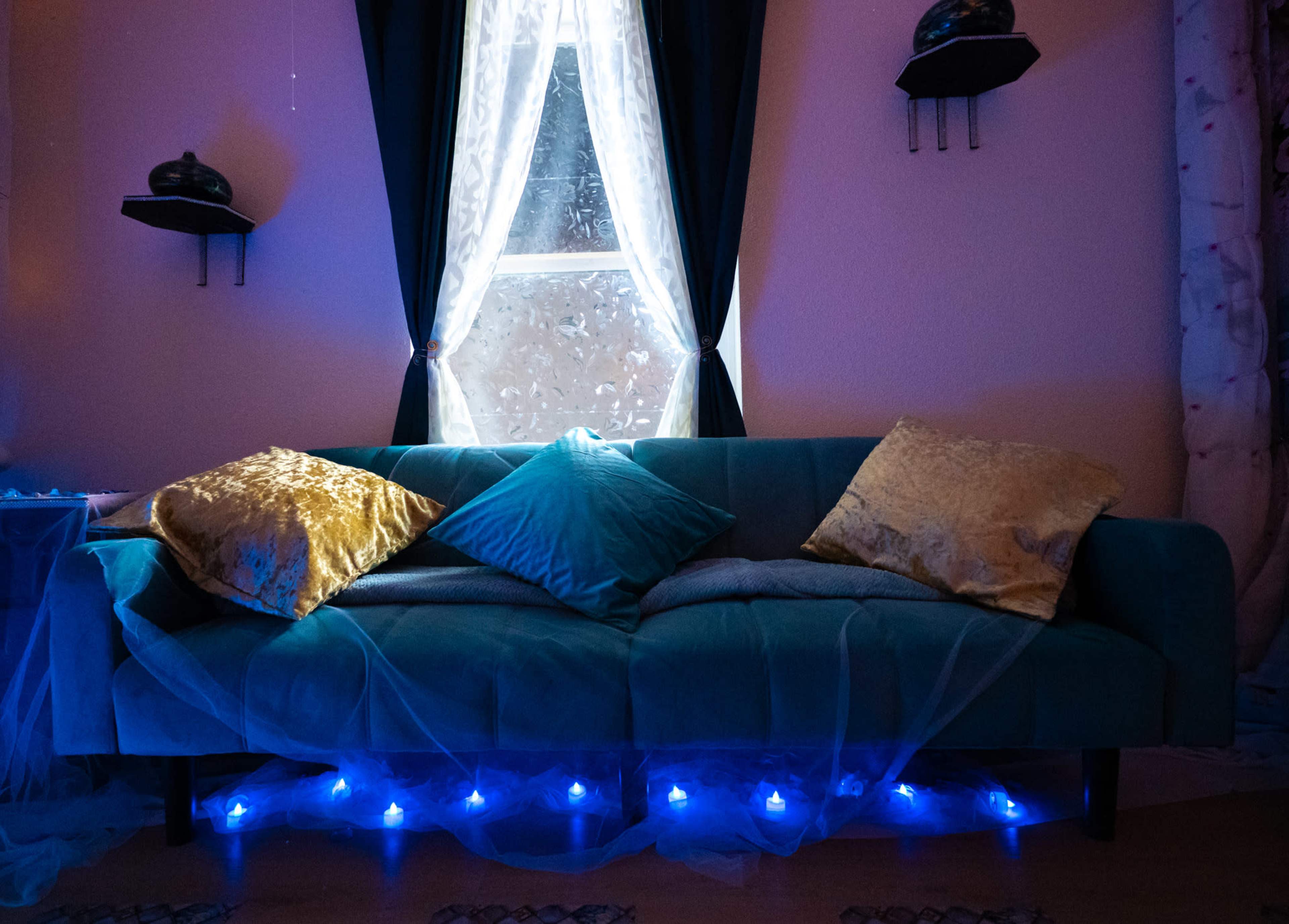 A teal couch adorned with decorative pillows sits in front of a window draped with sheer curtains, illuminated by blue lights.