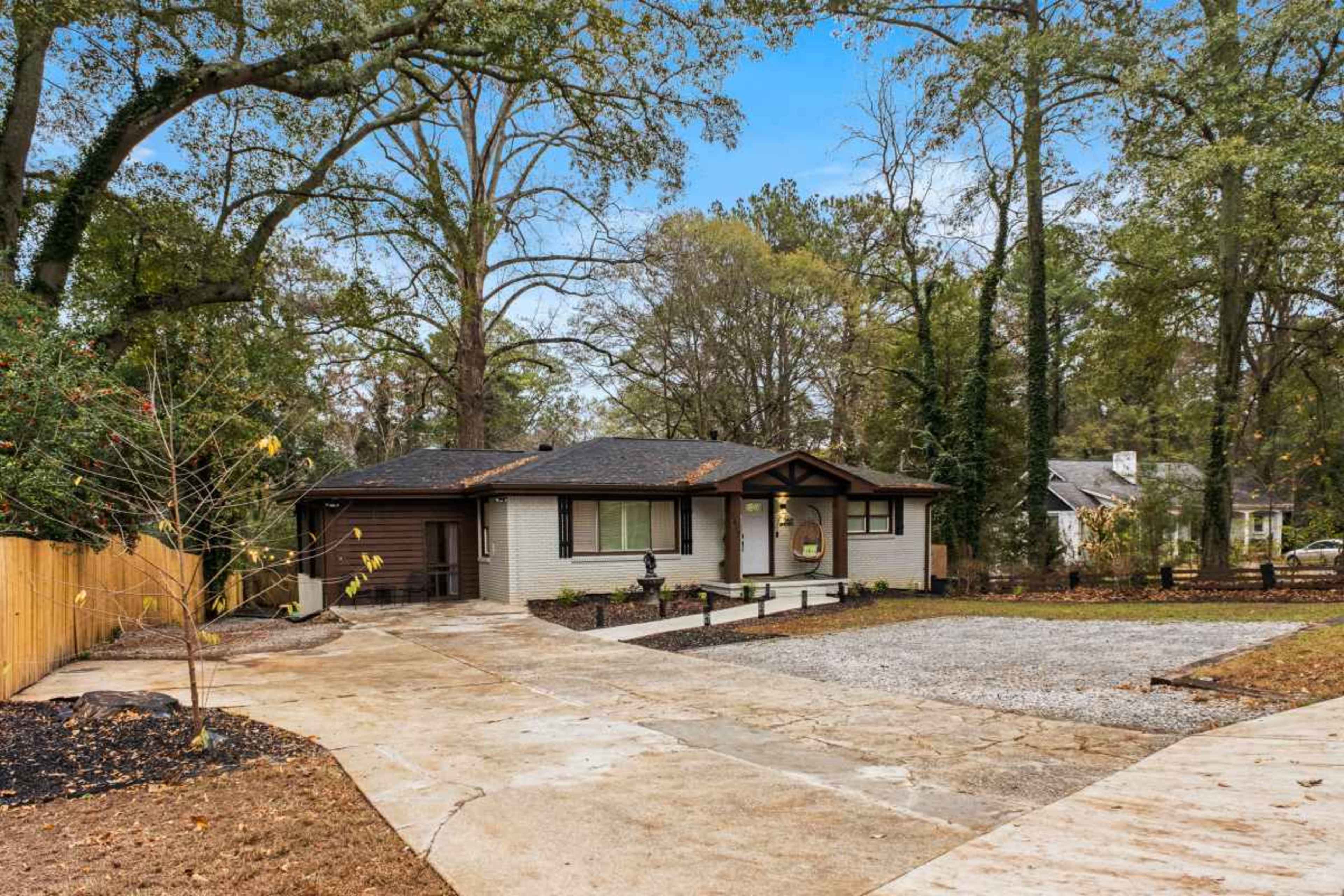 The image shows a single-story house with a dark roof surrounded by trees, featuring a gravel driveway and a landscaped yard.