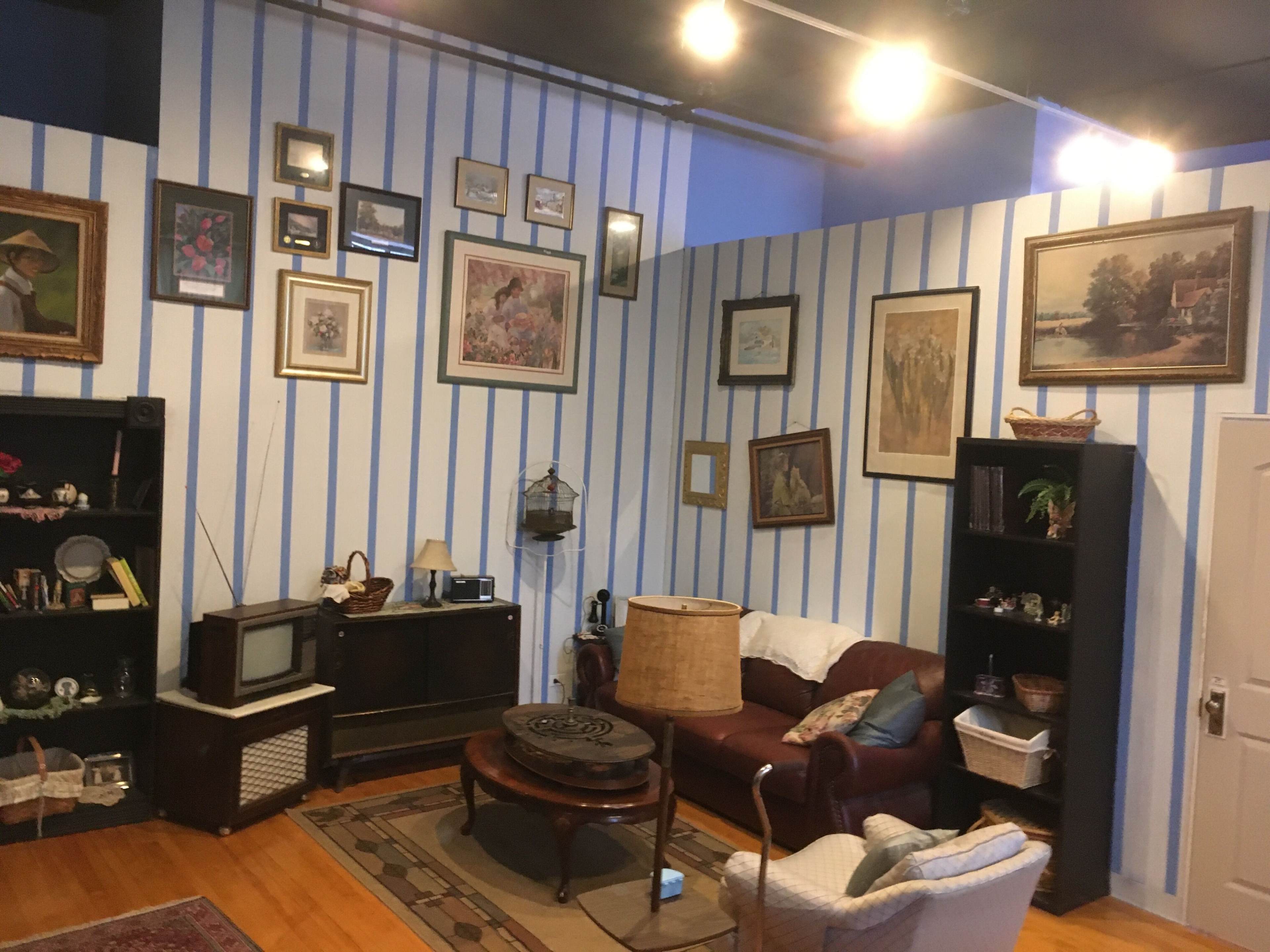 The room features striped walls adorned with various framed pictures, a vintage television set on a wooden cabinet, and a mix of upholstered furniture arranged around a central coffee table.