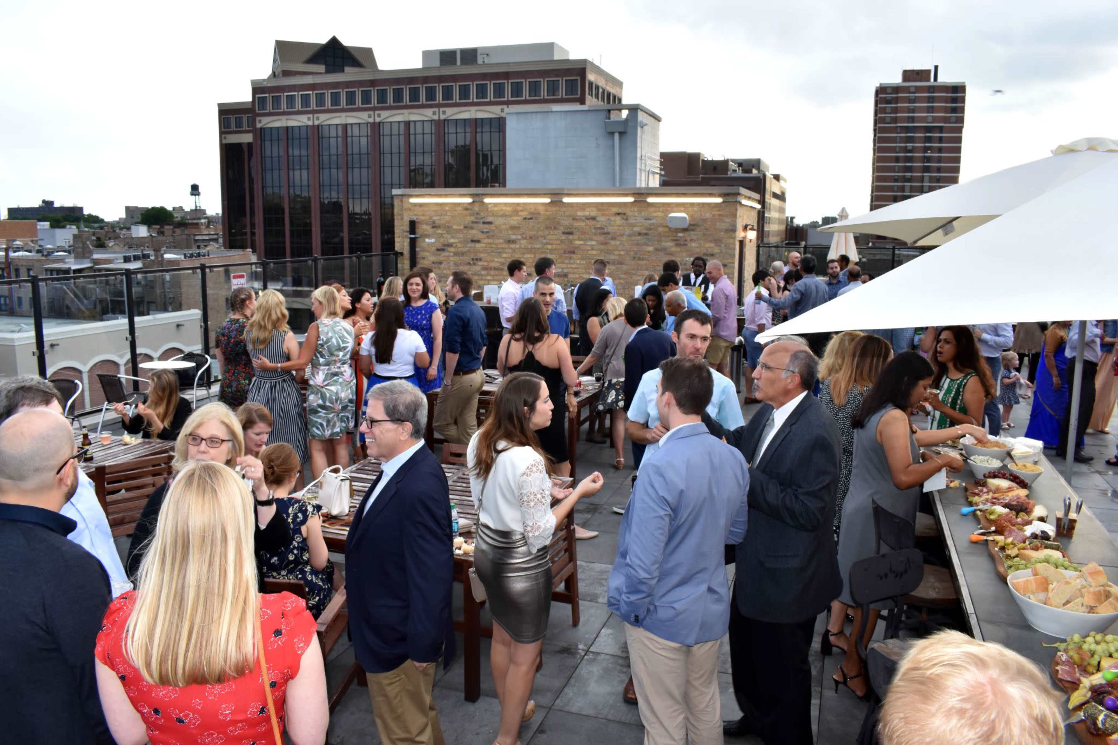 A large group of people mingles on a rooftop, with tables set up for food and drinks against a backdrop of urban buildings.