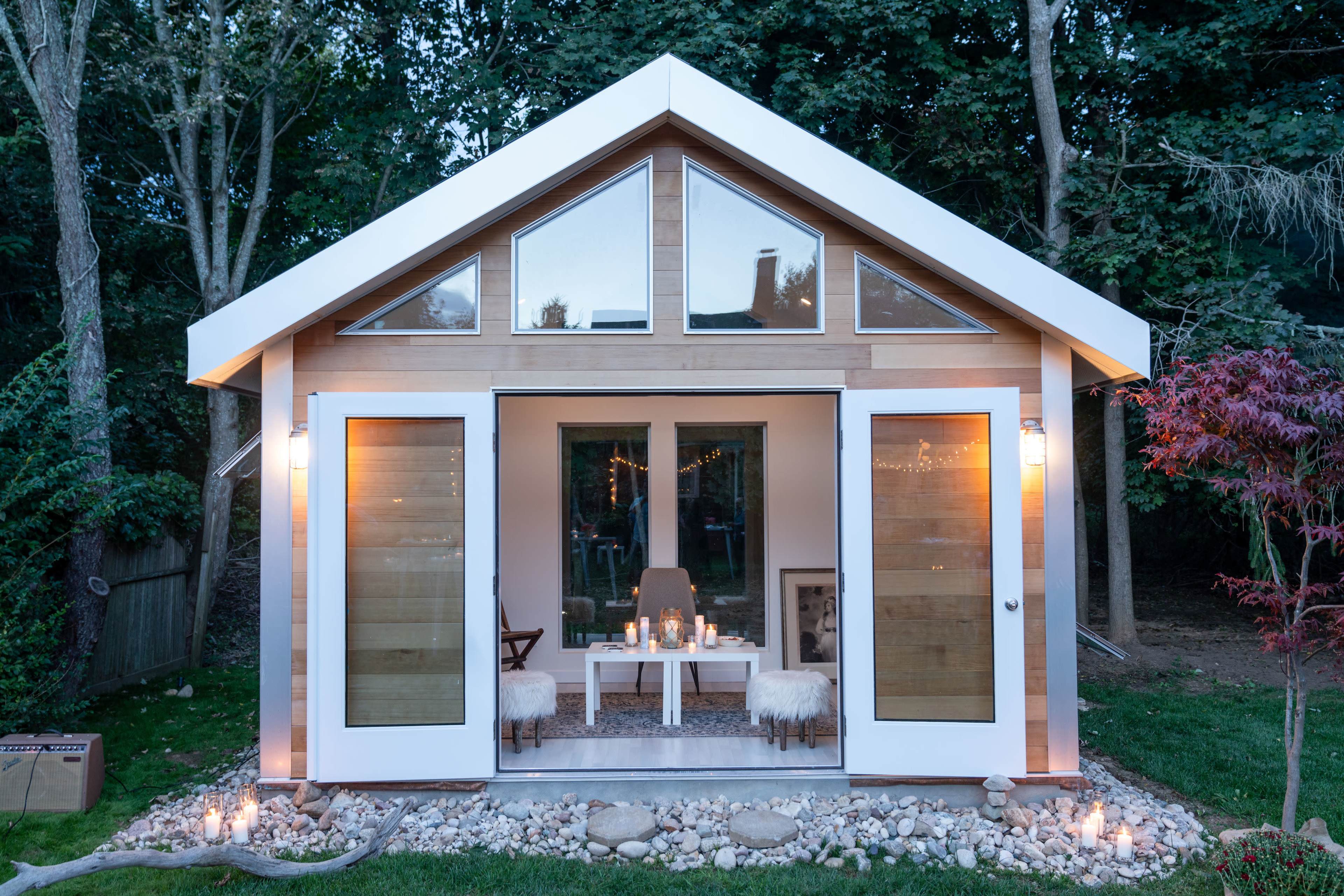 A small, modern cabin with large windows and a wooden exterior is surrounded by landscaping featuring a rock garden and soft outdoor lighting.