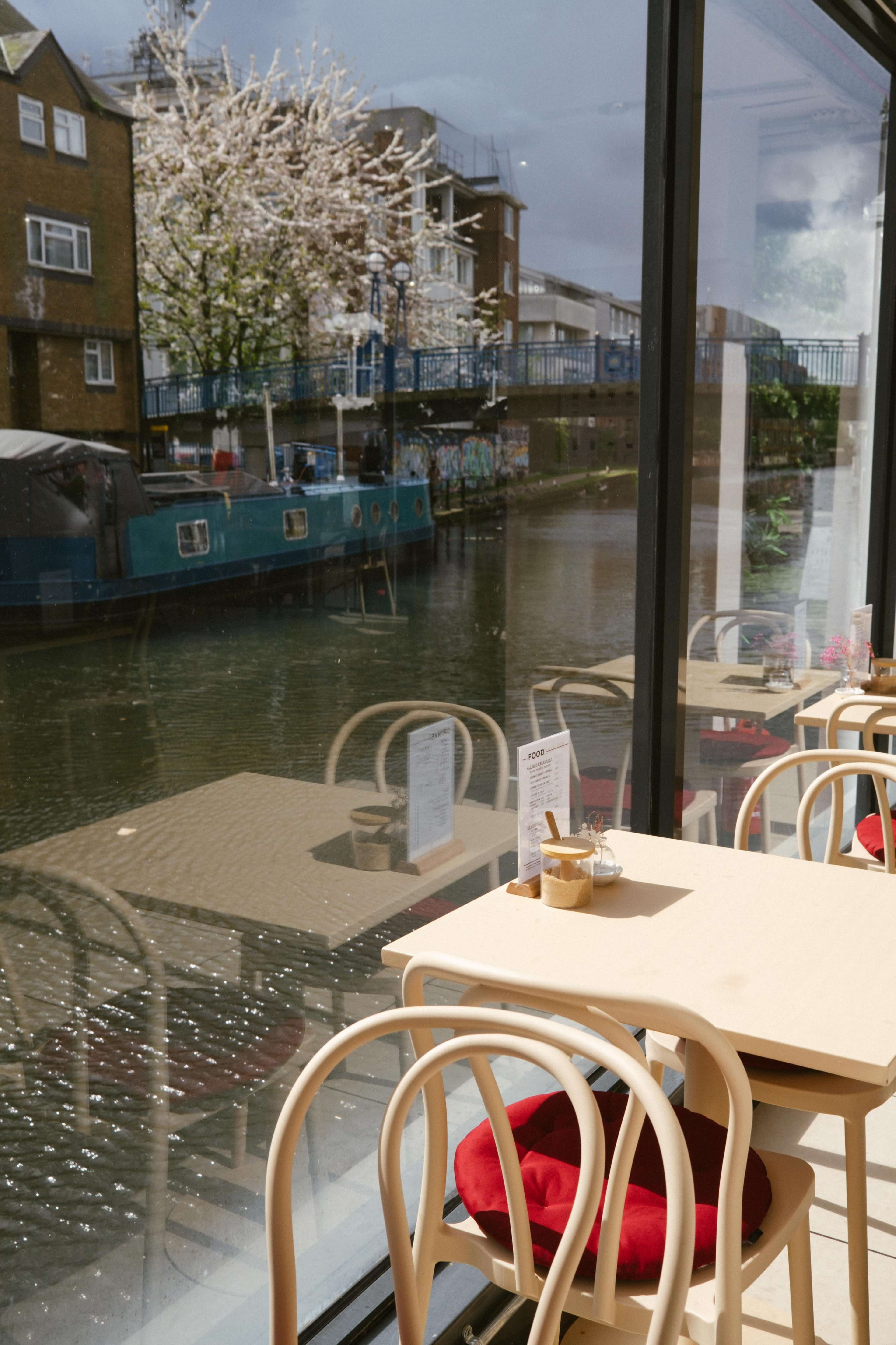A café with wooden tables and chairs inside, overlooking a canal with a blue boat and trees outside.