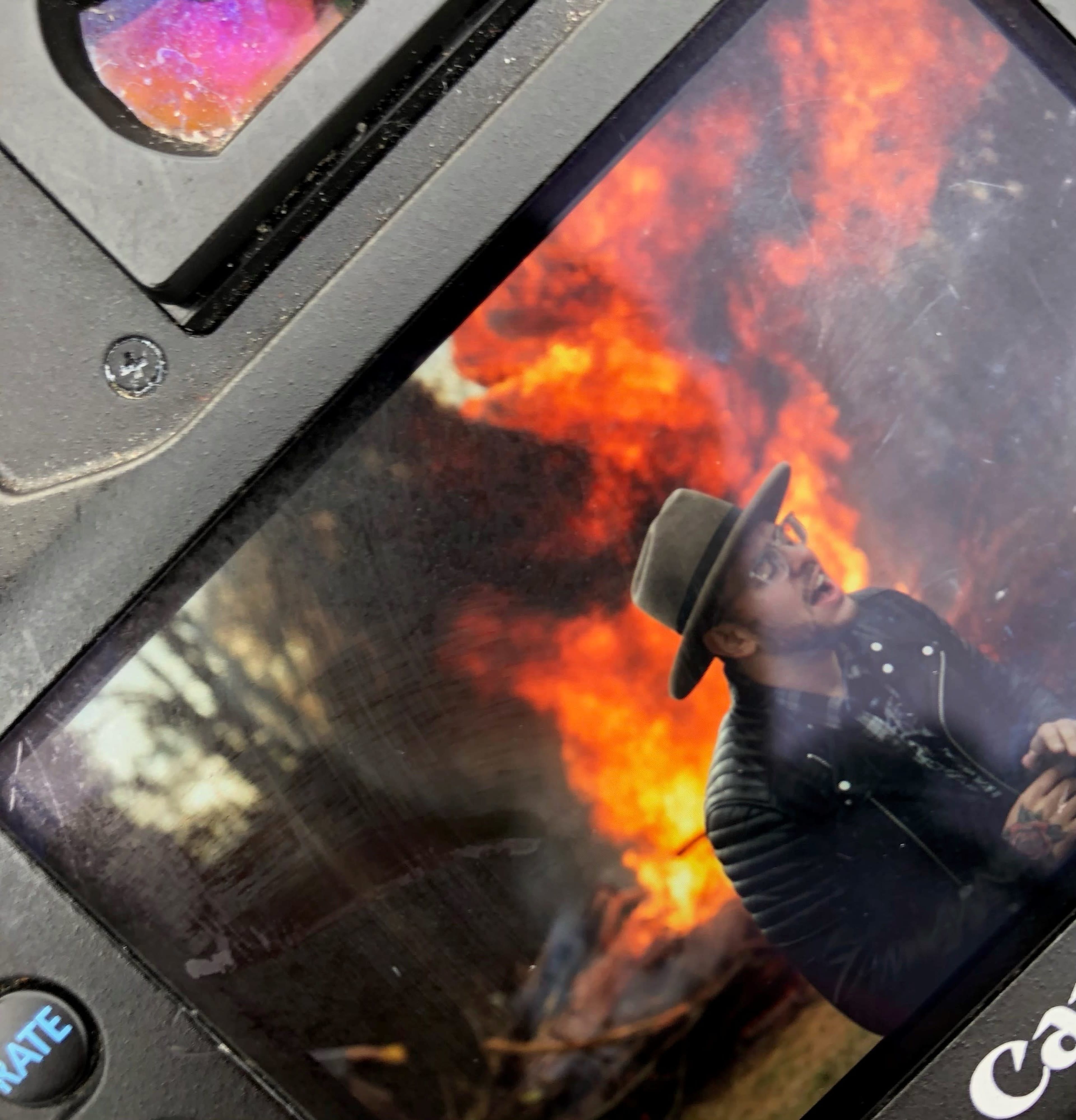 A man wearing a hat and a jacket is seen in front of a large fire.
