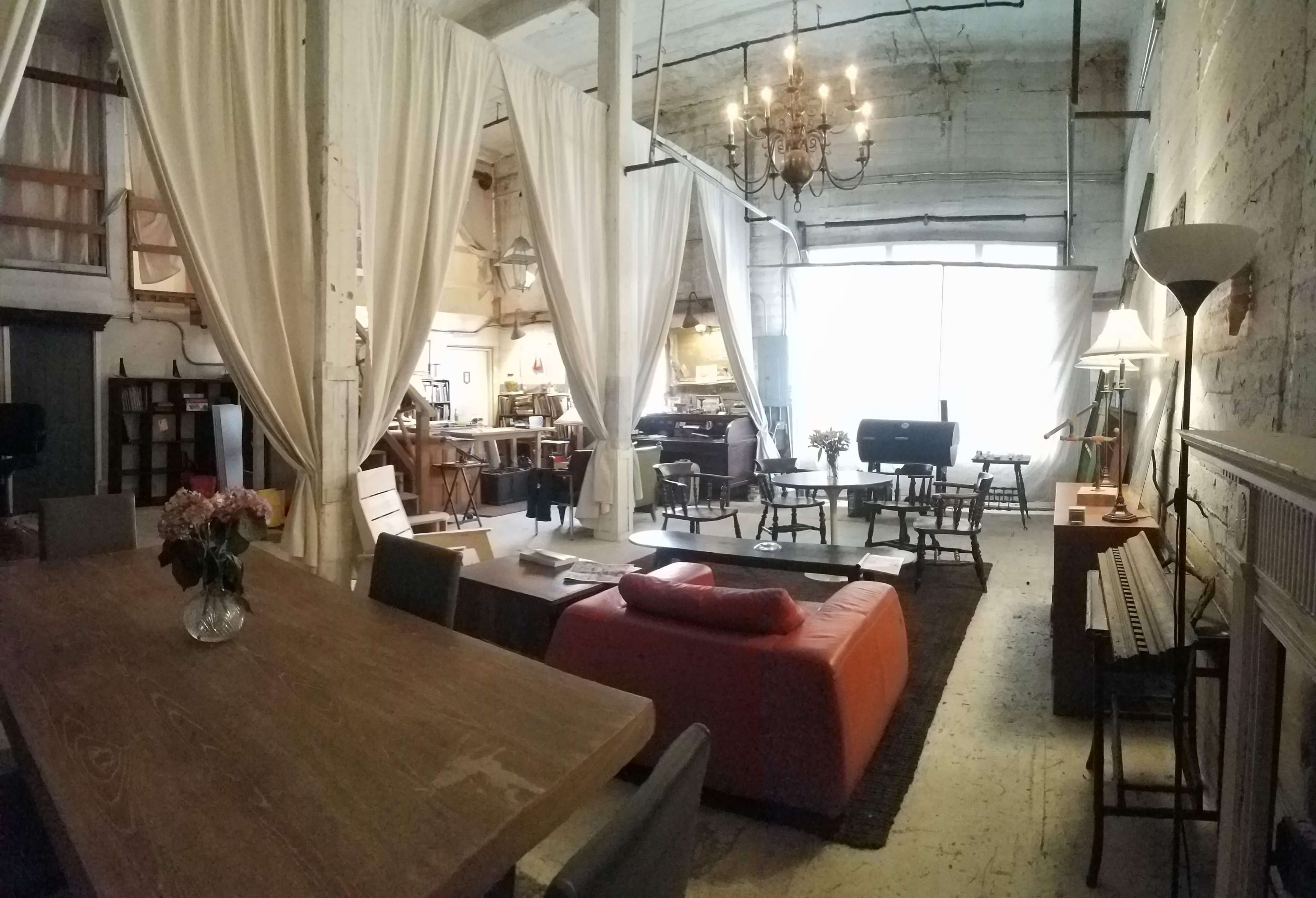 A spacious loft-style interior with a dining area, living space, and multiple seating arrangements, separated by fabric drapes and illuminated by a chandelier.