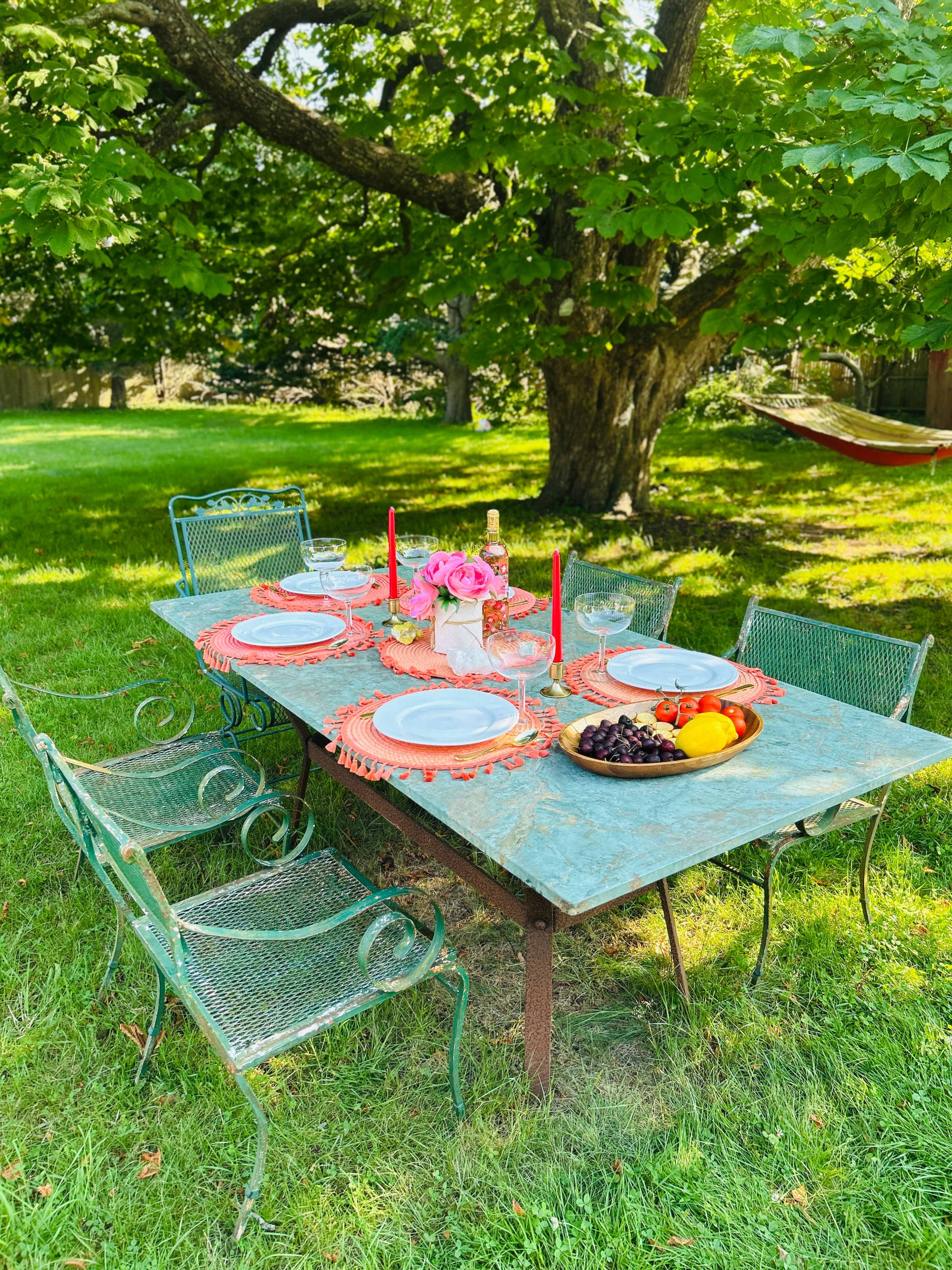 A rustic outdoor dining table is set with dinnerware, glasses, fruit, and candles beneath a leafy tree in a grassy area.