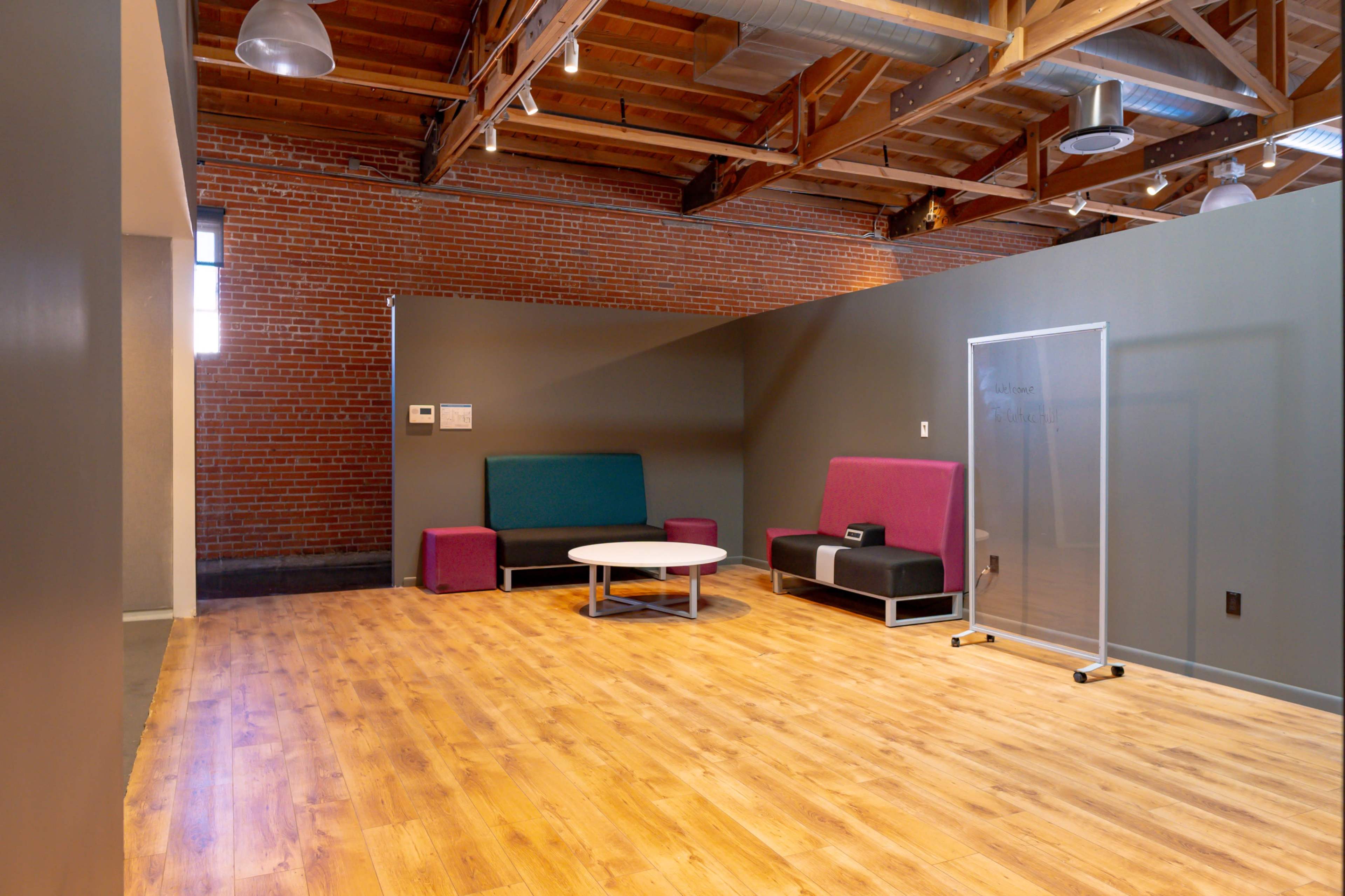 A spacious, minimalist room with wooden flooring features a circular table and two contrasting sofas against a grey wall, with exposed beams overhead.