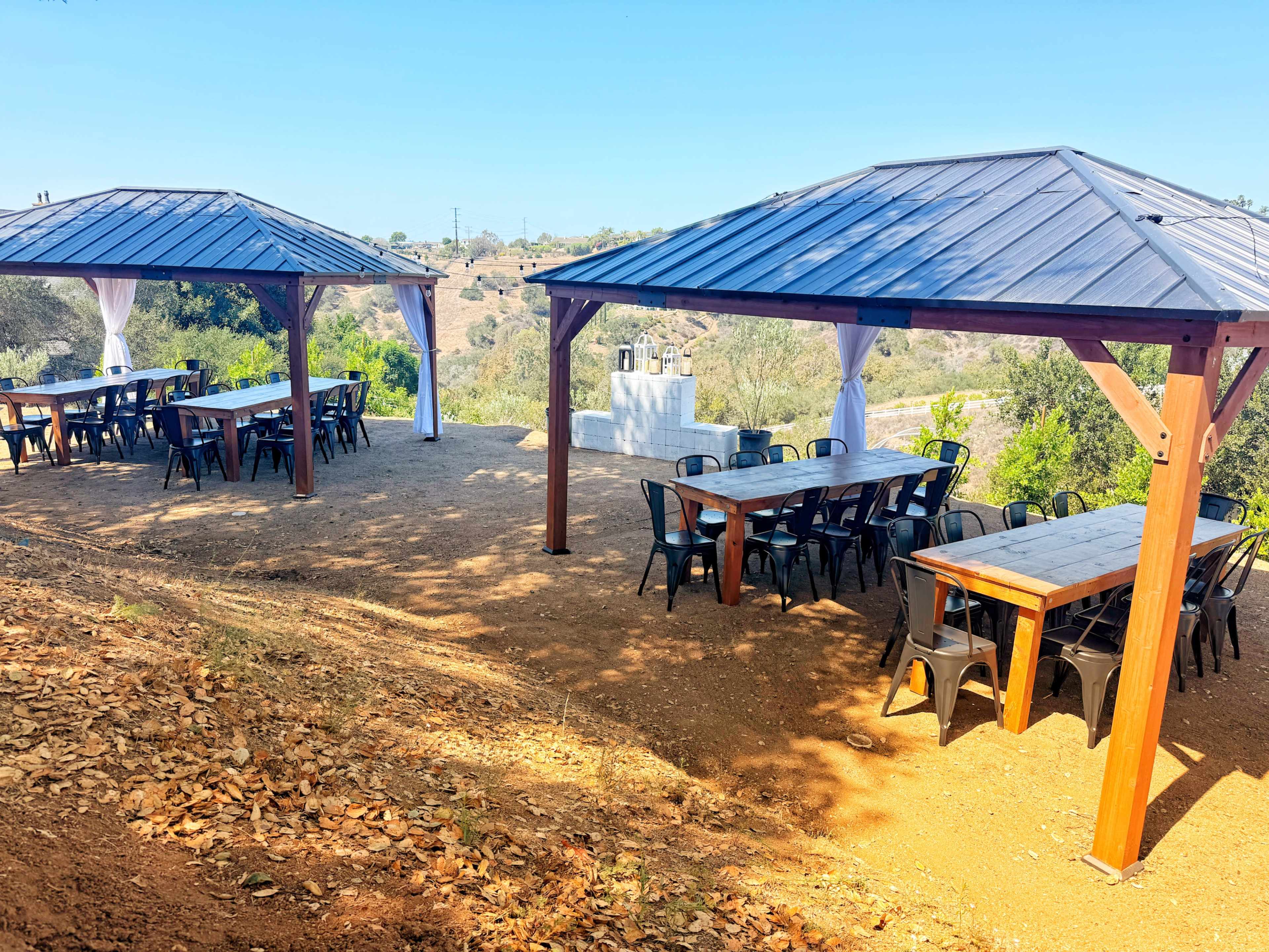The image shows two open-air pavilions with wooden tables and black chairs set on a dirt area, surrounded by greenery and hills under a clear blue sky.