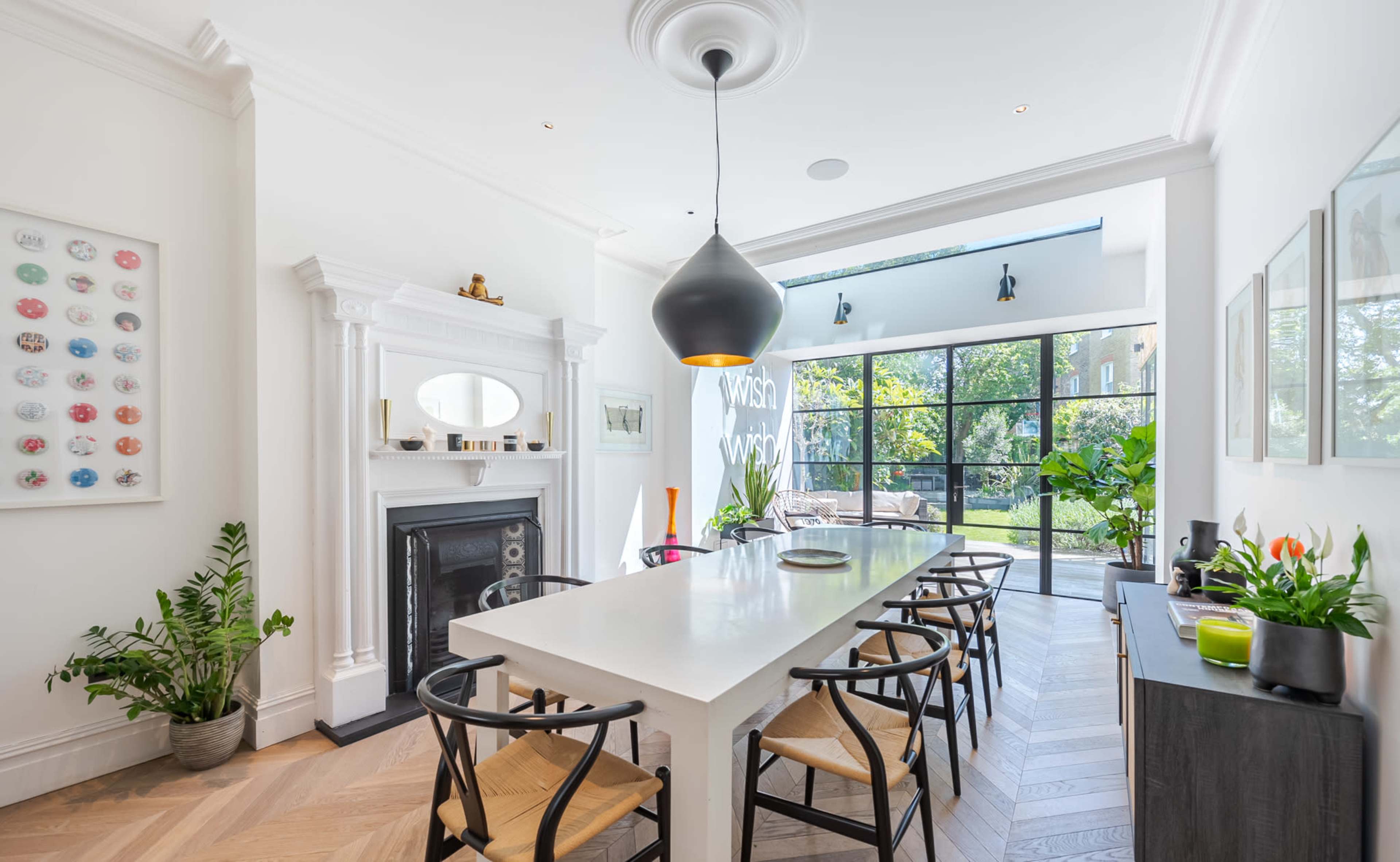 A spacious dining area features a long white table surrounded by black chairs, with a large window overlooking a garden and a modern light fixture hanging above.