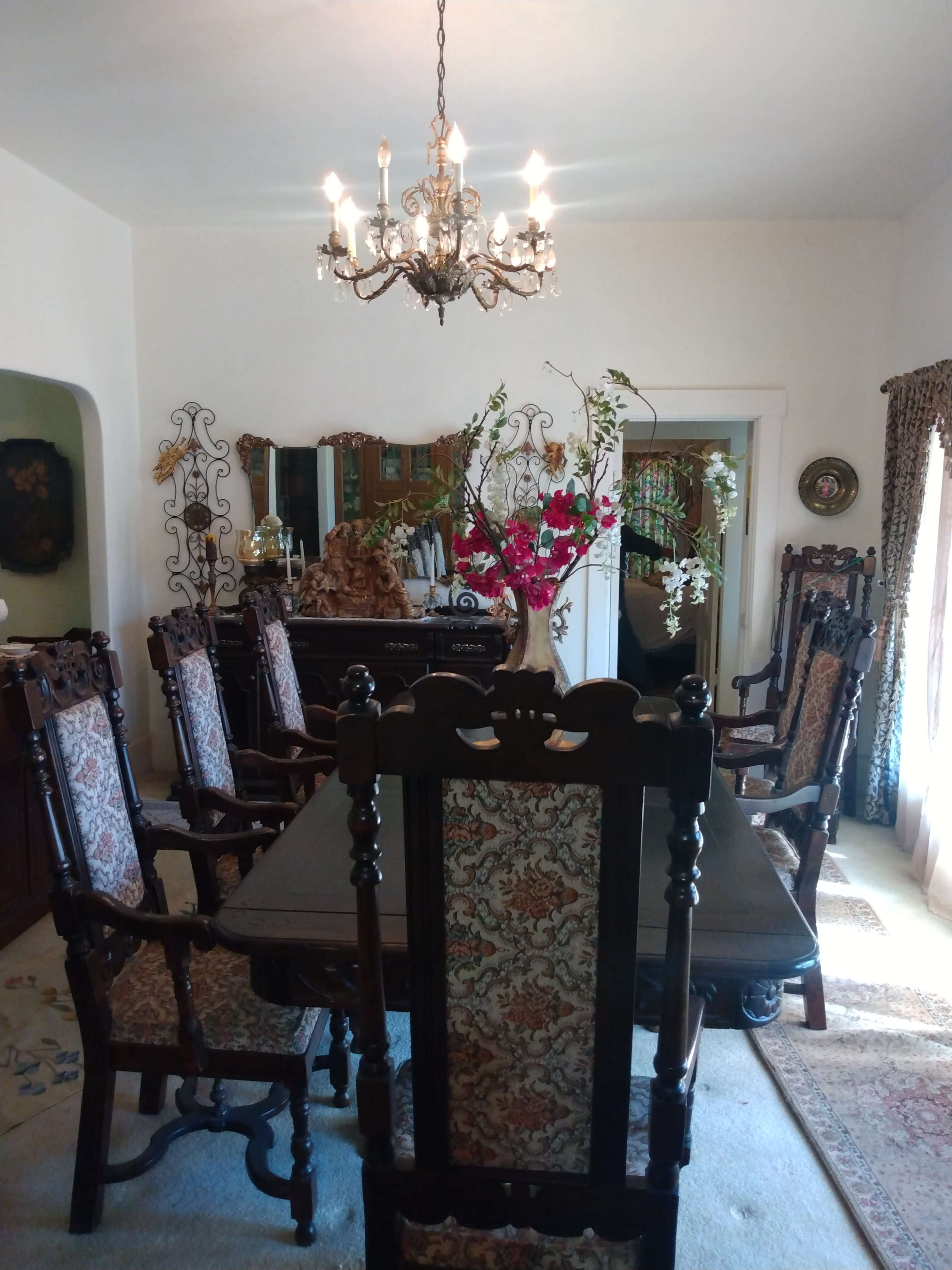 A dining room with a long table surrounded by ornate wooden chairs, a chandelier overhead, and decorative floral arrangements.