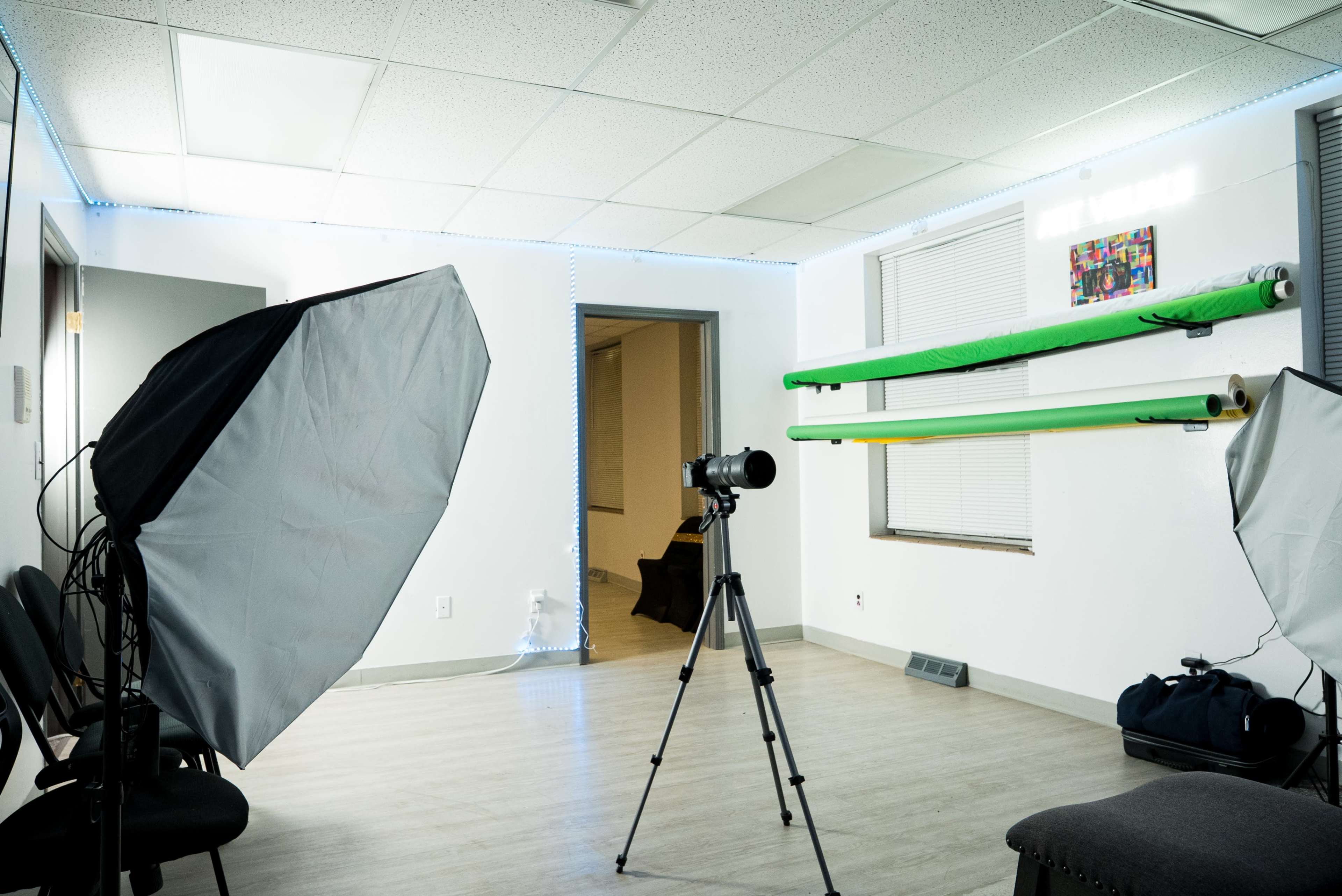 The image shows a photography studio with two softbox lights, a tripod with a camera, and rolls of green seamless backdrop paper mounted on a wall.
