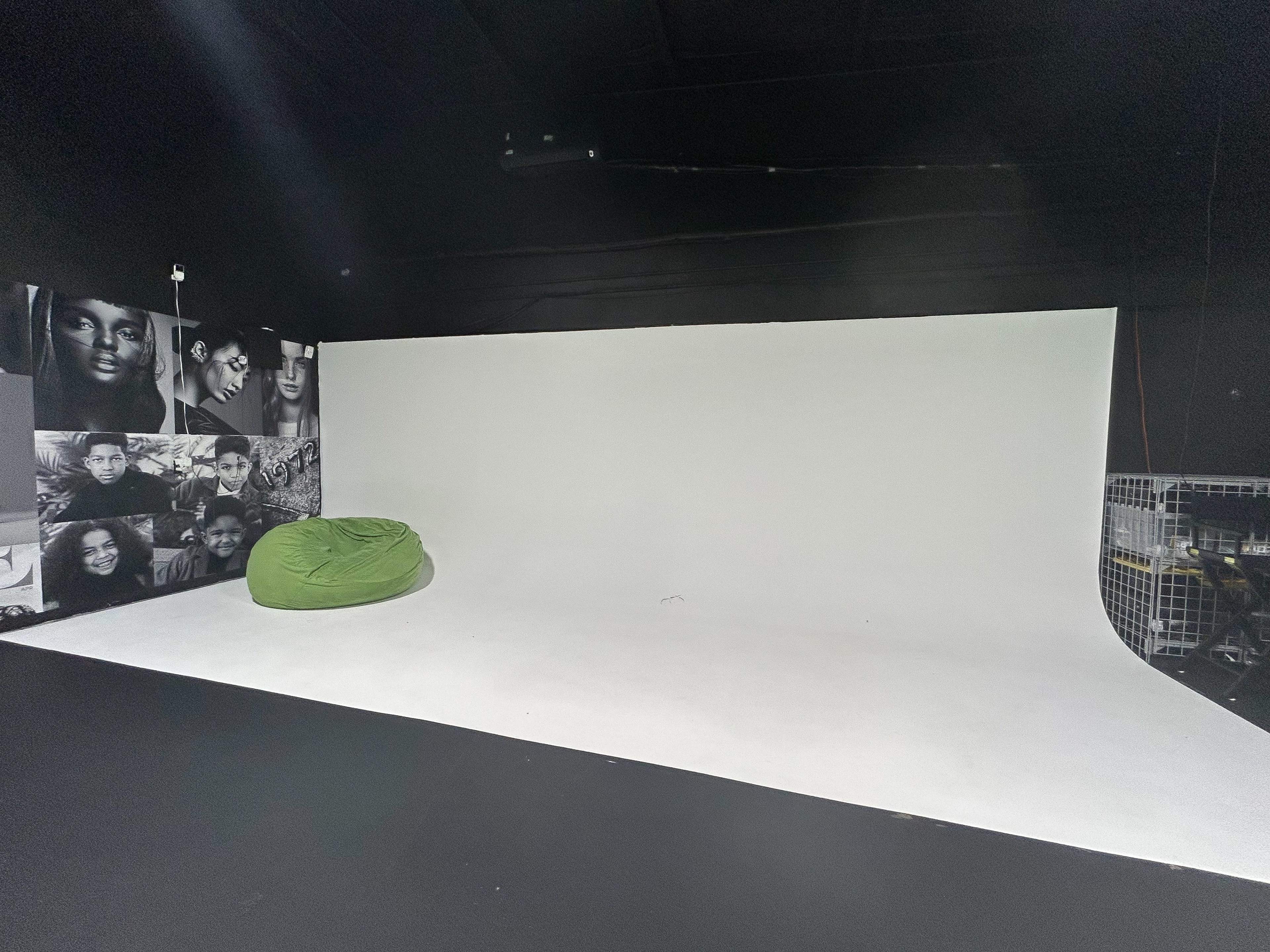 A studio space with a white backdrop, a green beanbag chair, and black walls featuring black-and-white portraits.