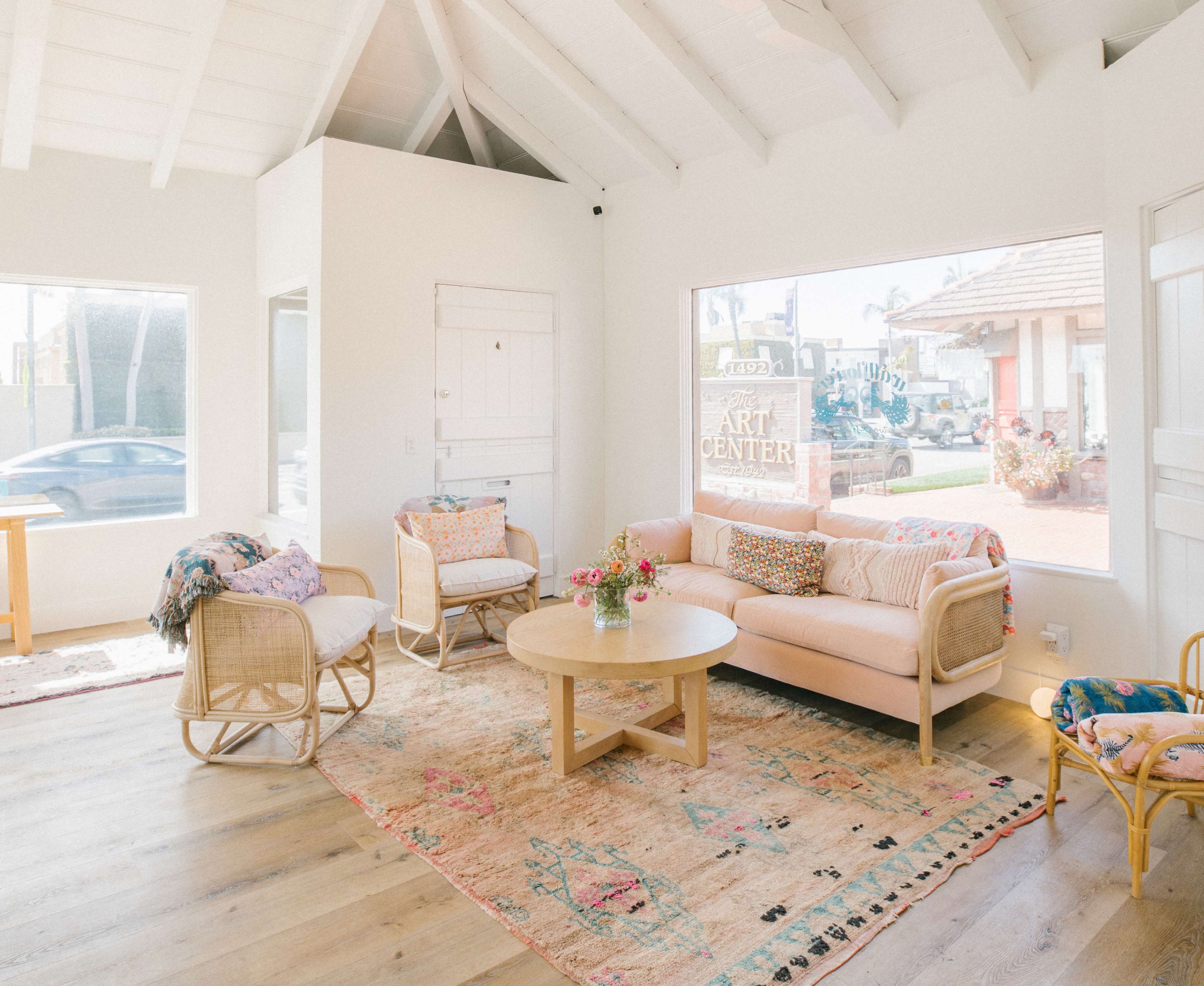 A bright, airy living room features a pink sofa, a round table, and rattan chairs arranged on a colorful rug, with large windows allowing natural light to fill the space.