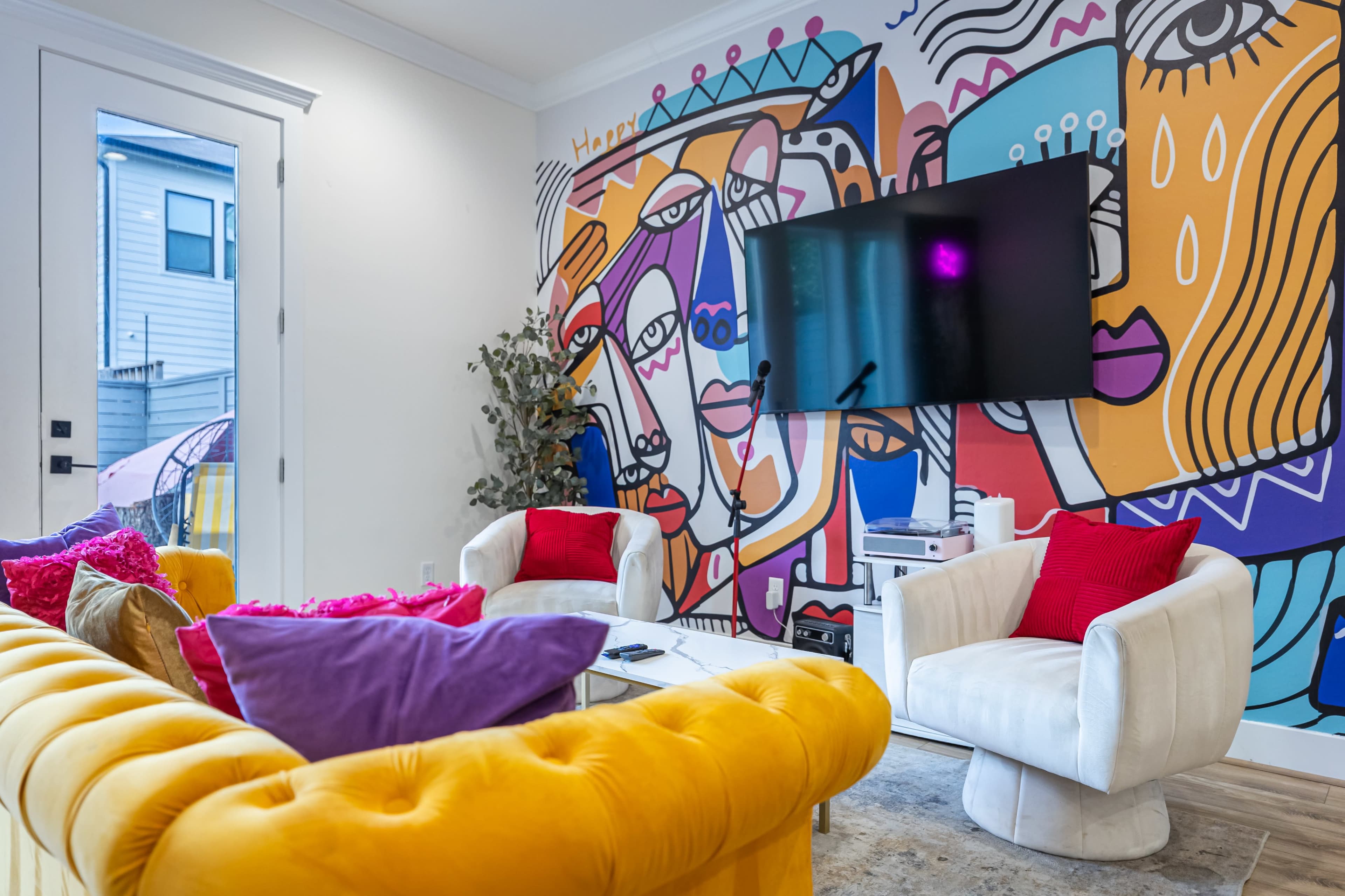 The image depicts a modern living room with a colorful mural on the wall, featuring abstract faces, and two white armchairs positioned around a glass coffee table.