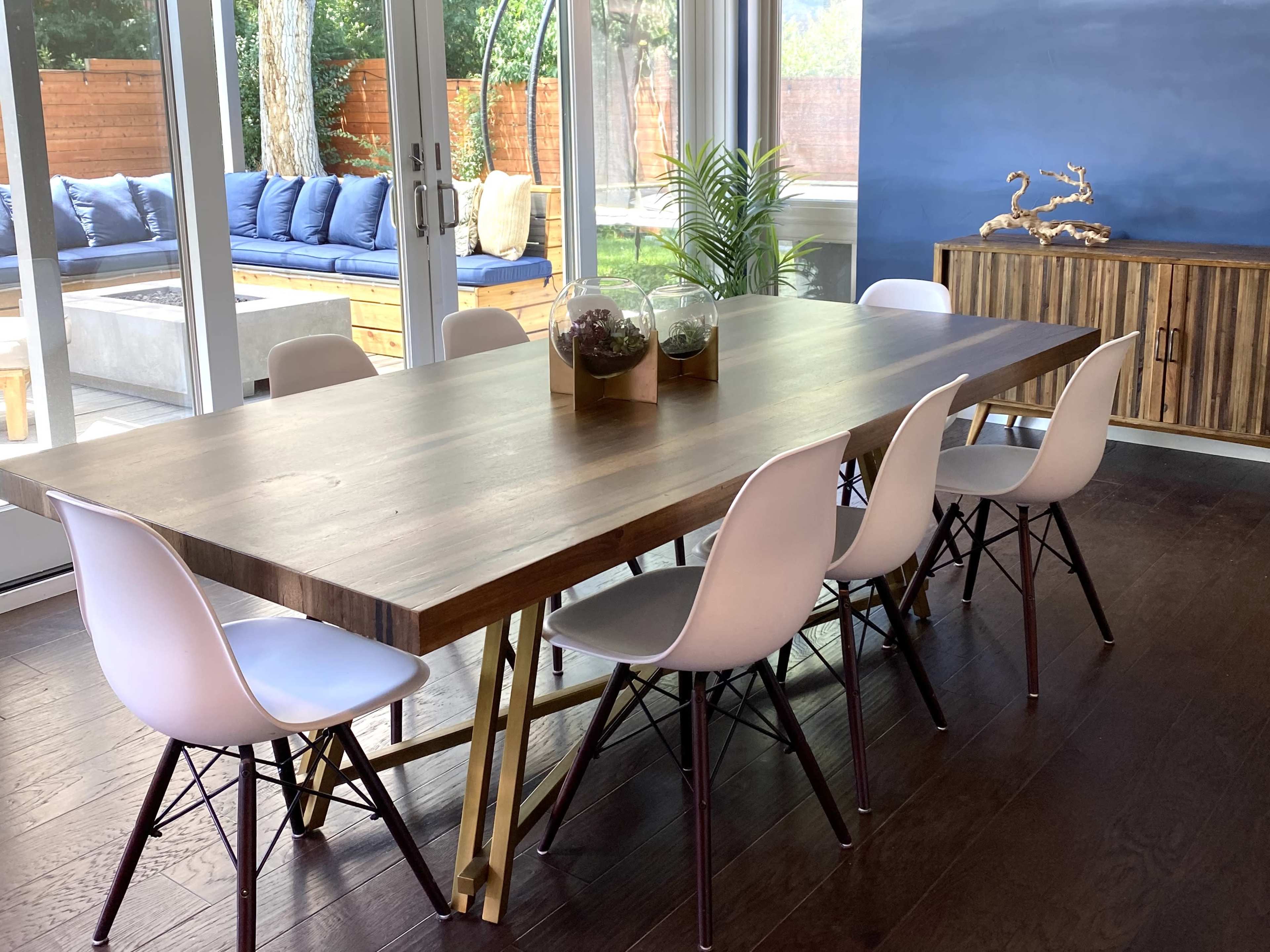 A wooden dining table surrounded by six white chairs is set in a modern room with large windows and a view of an outdoor seating area.