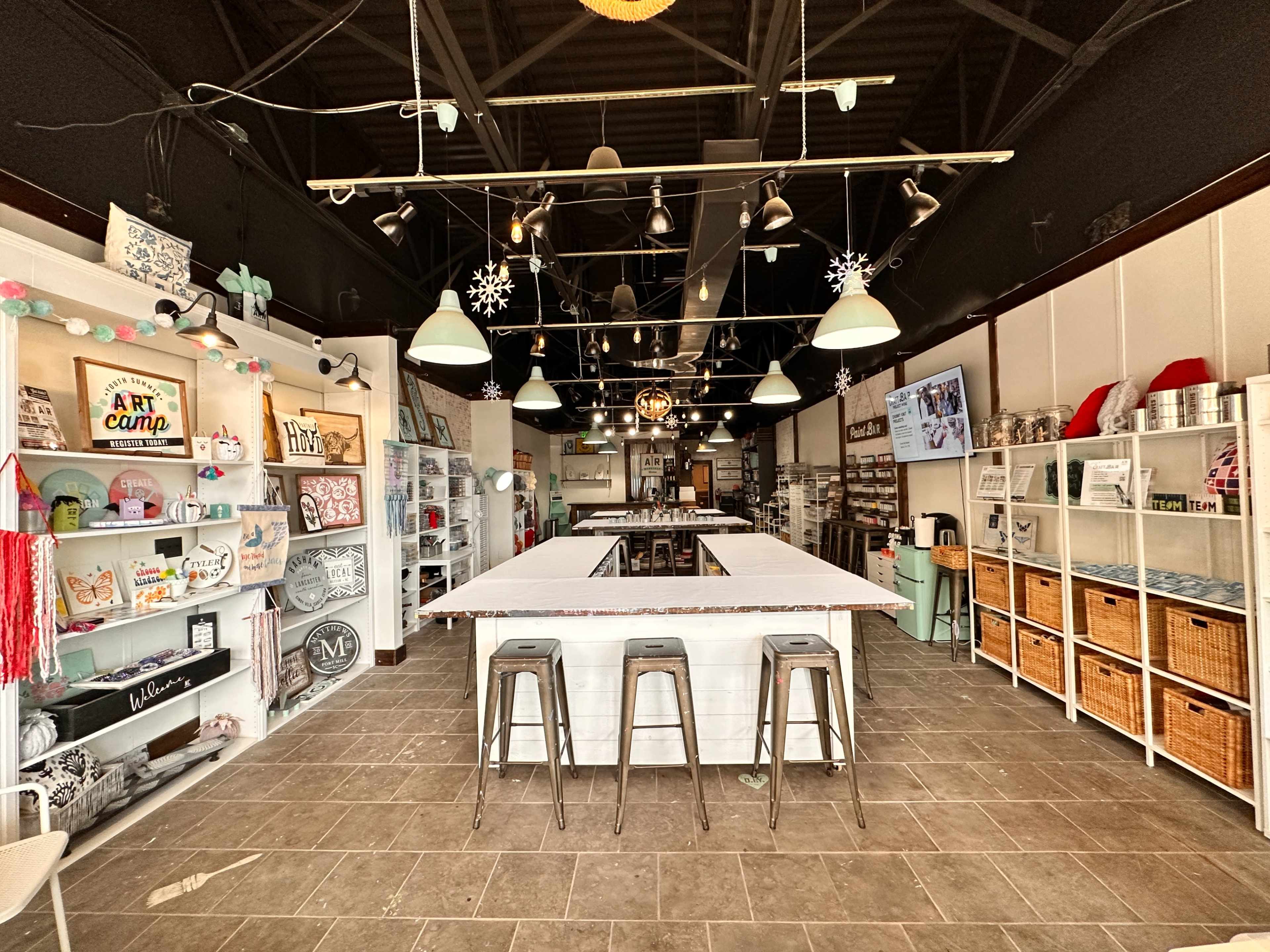 The image shows a craft store interior featuring a central work table, shelving with various art supplies, and a light fixture with pendant lamps.