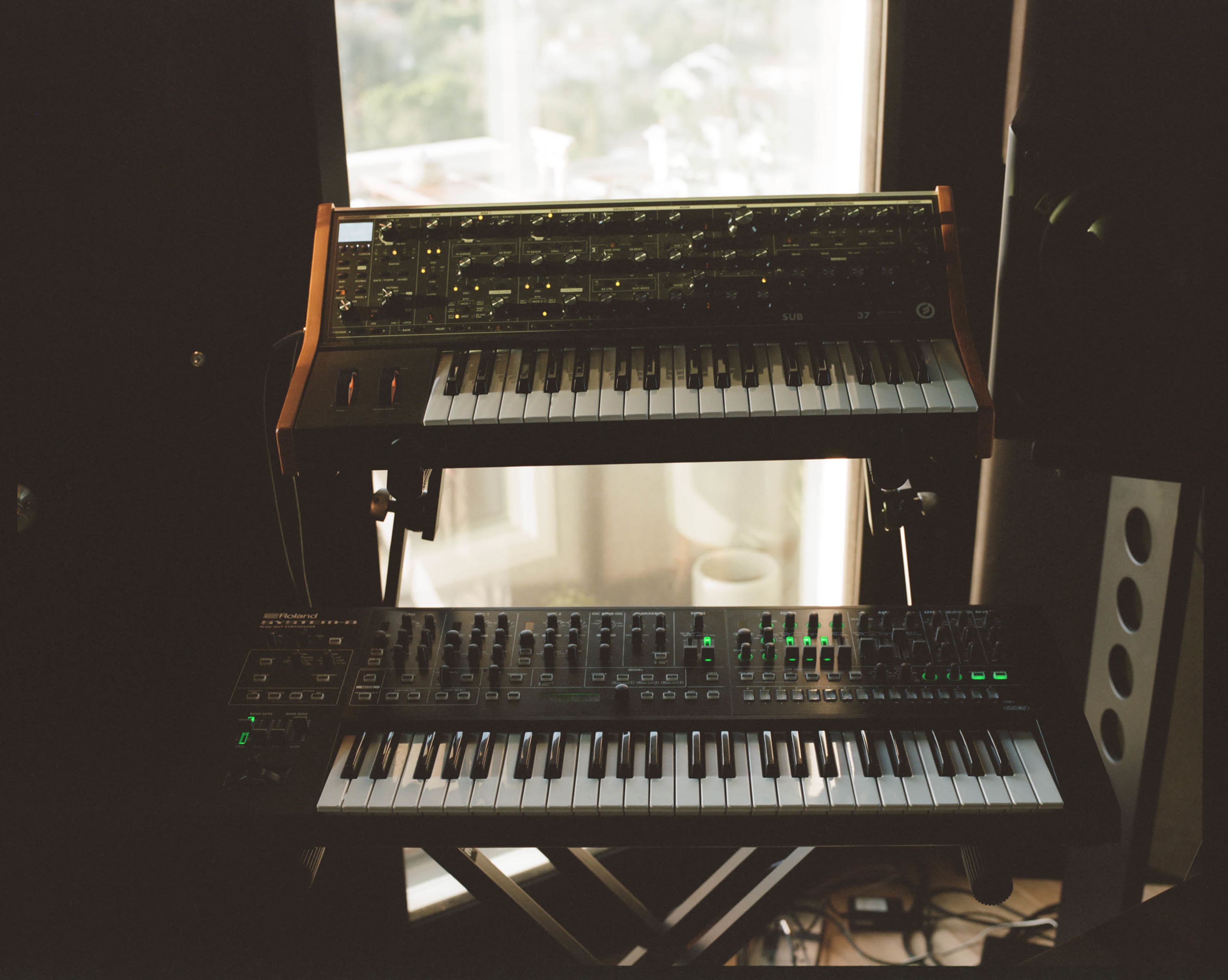 The image shows two synthesizers with keyboards arranged on a stand in a music studio near a window.